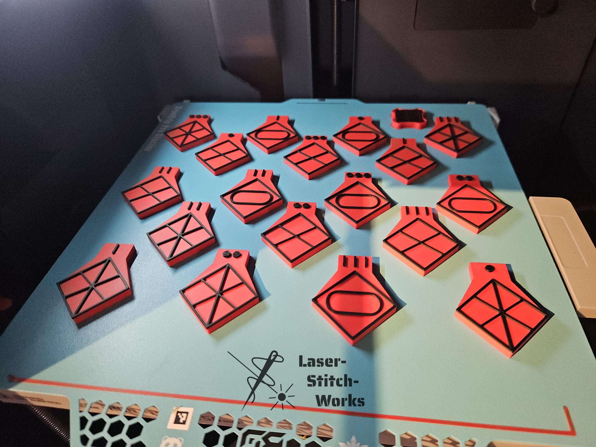 Mehrere rote und schwarze lasergravierte Holzstücke in zeichenförmigen Formen, die auf einem blauen Tisch mit dem Logo und dem Text 'Laser-Stitch-Works' liegen.