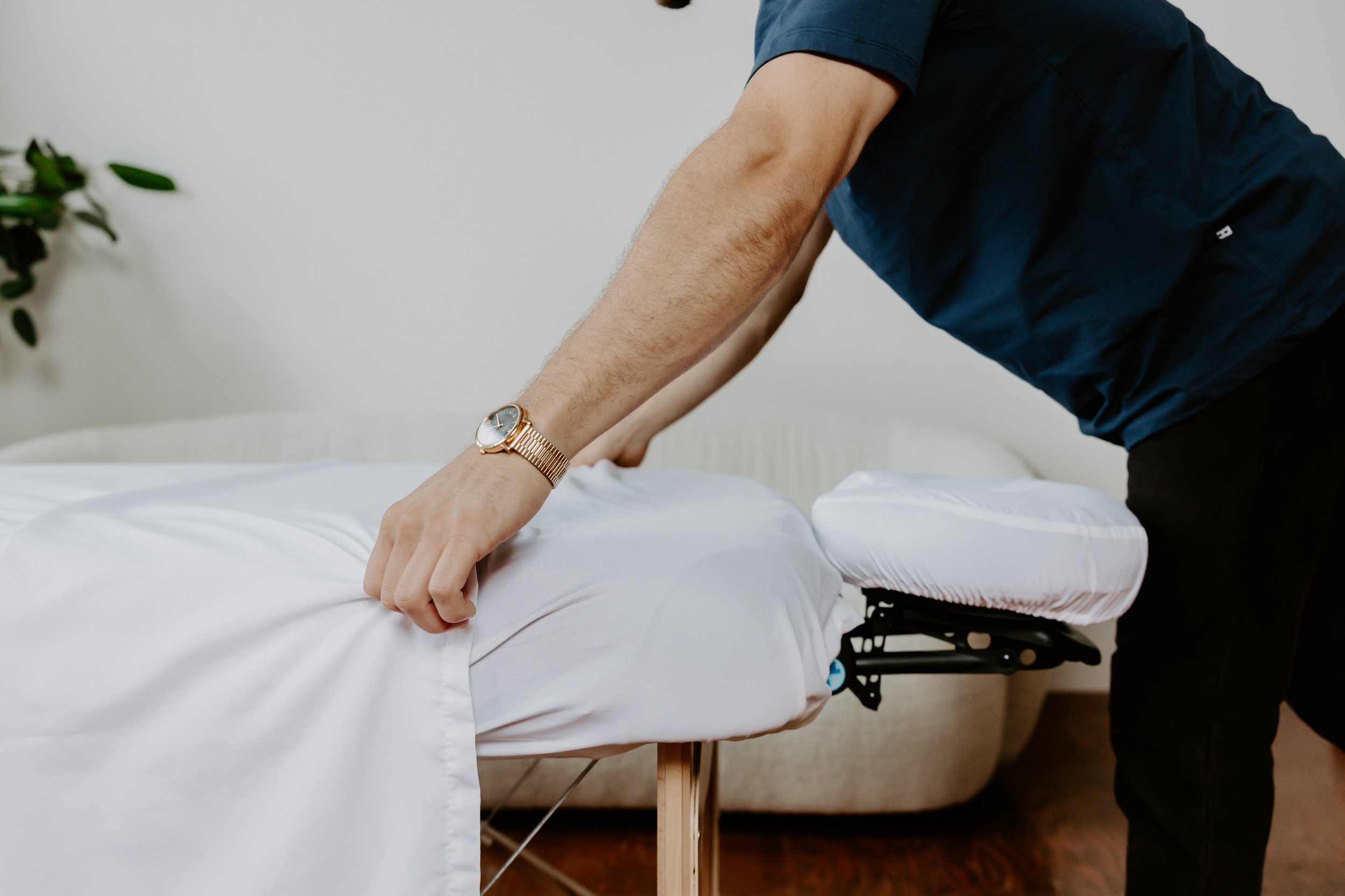 A person giving a massage or performing therapy on a client lying face down on a massage table, with a white sheet covering the client and a green plant partially visible in the background.