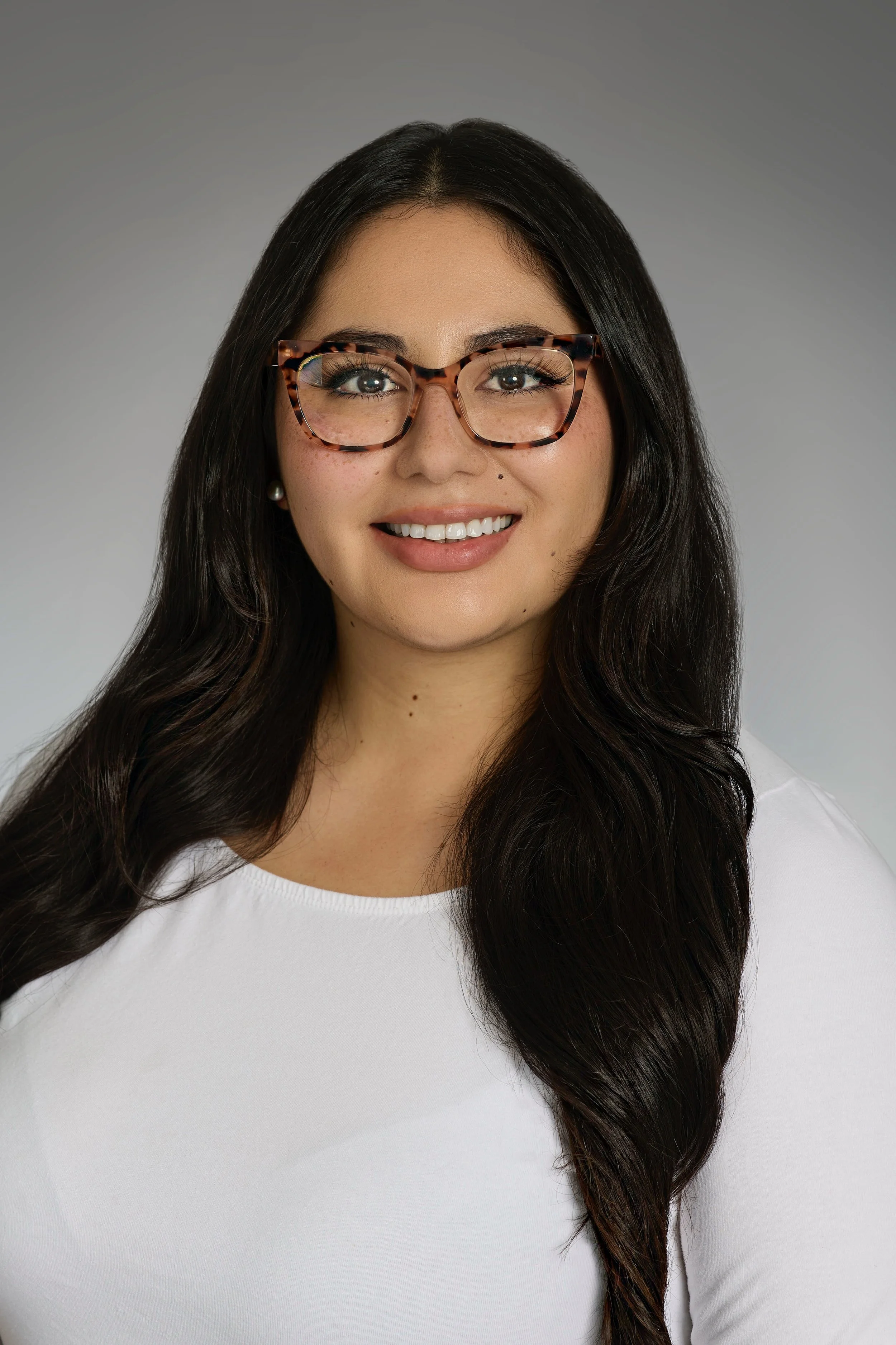 Portrait of a woman with long dark hair, wearing tortoiseshell glasses and a white top, smiling against a gray background.