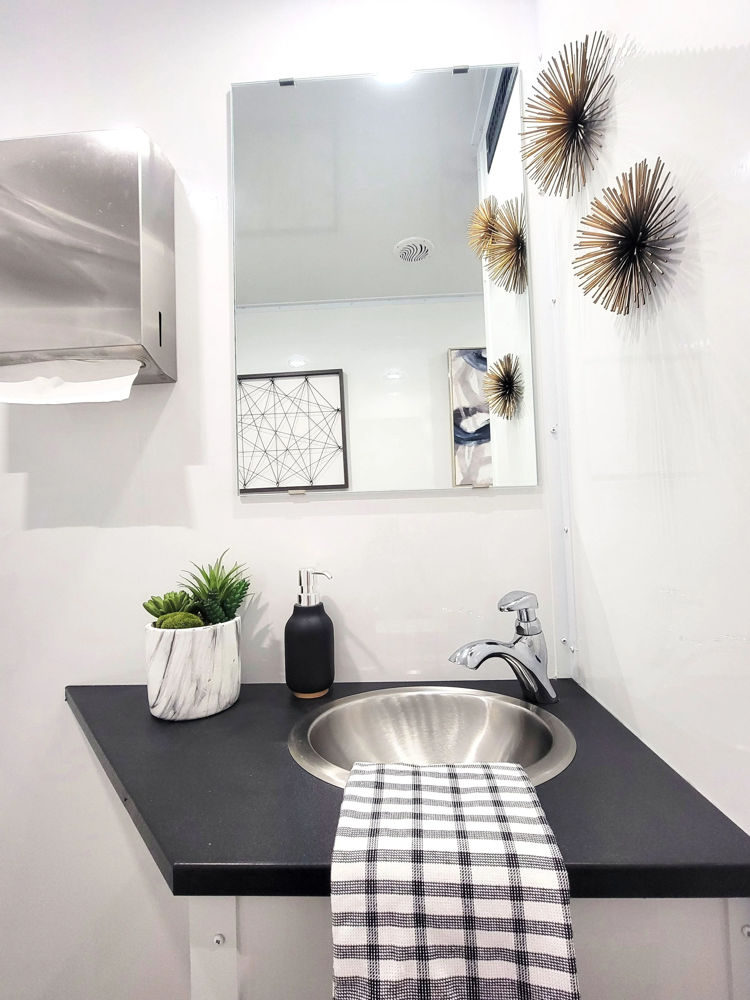 A bathroom sink with a mirror above, decorated with a small plant in a white marble pot and a black soap dispenser, and a checkered dish towel hanging over the black countertop, with decorative wall art and metallic starburst wall sculptures.