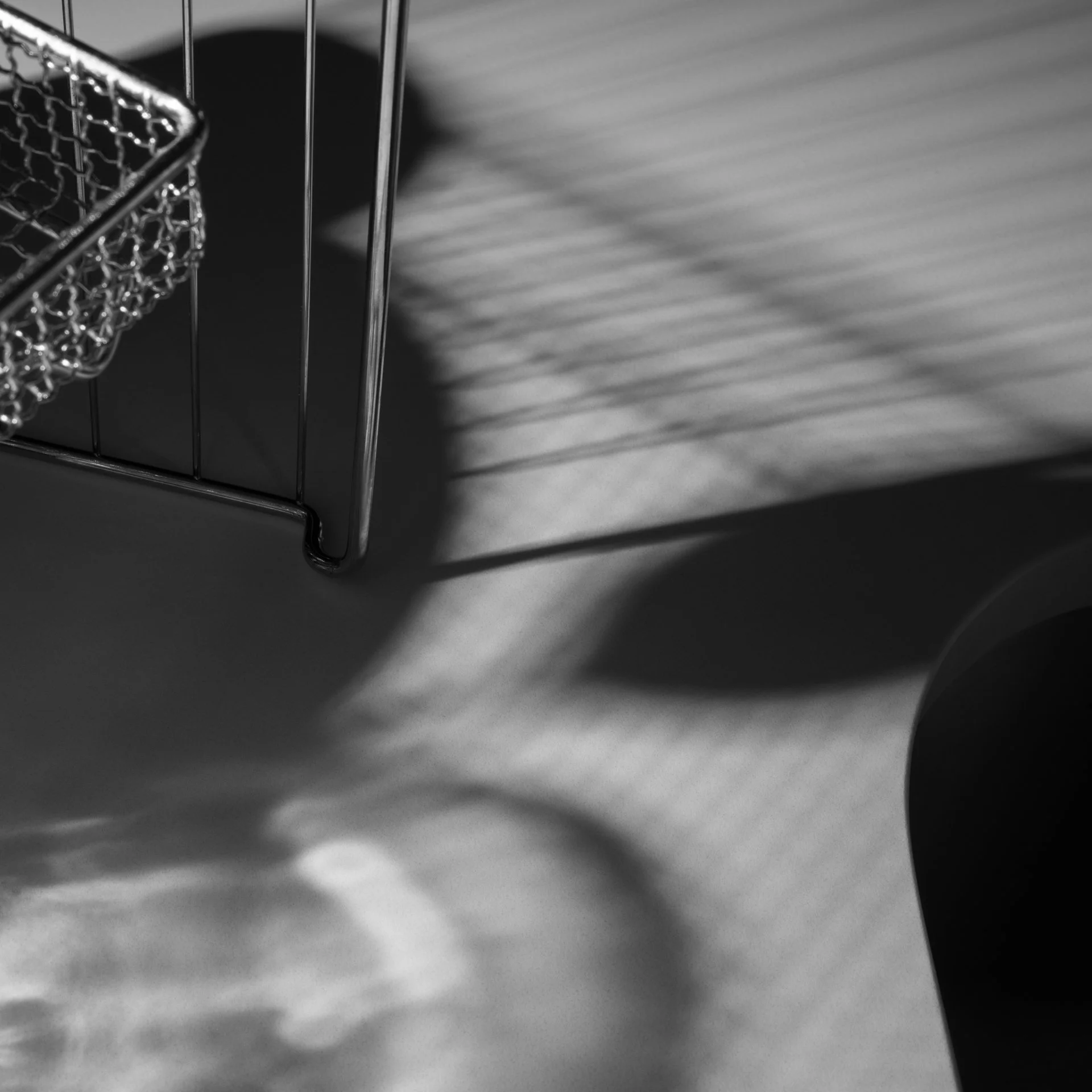  Black and white photo of metal wire basket casting geometric shadows on a surface, with overlapping patterns created by light and basket, and part of a dark, rounded object in the lower right corner. 