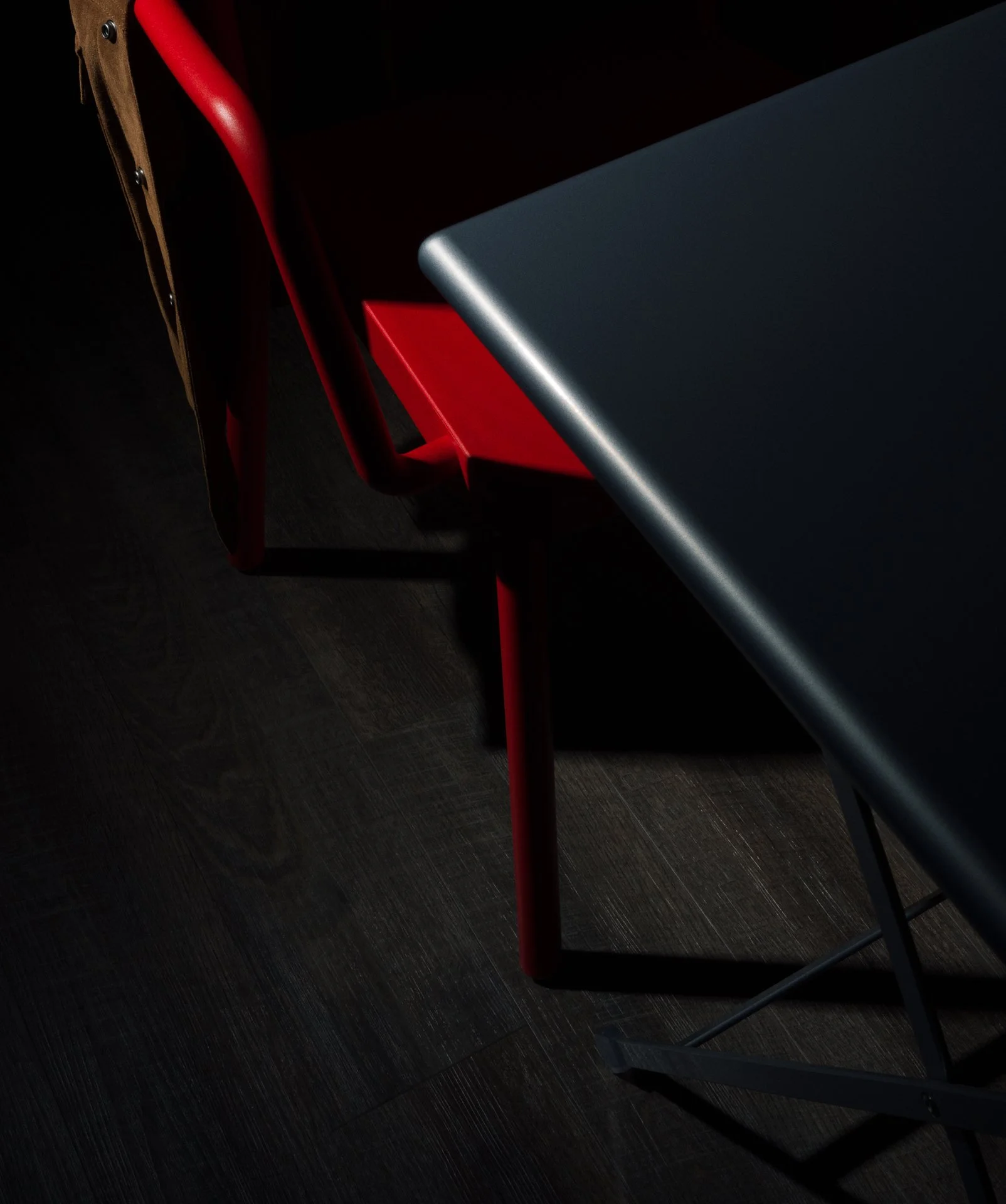  A dark, moody photo showing a dark silver metal table with sharp edges and a red plastic chair underneath, partially illuminated, on a dark wooden floor. 