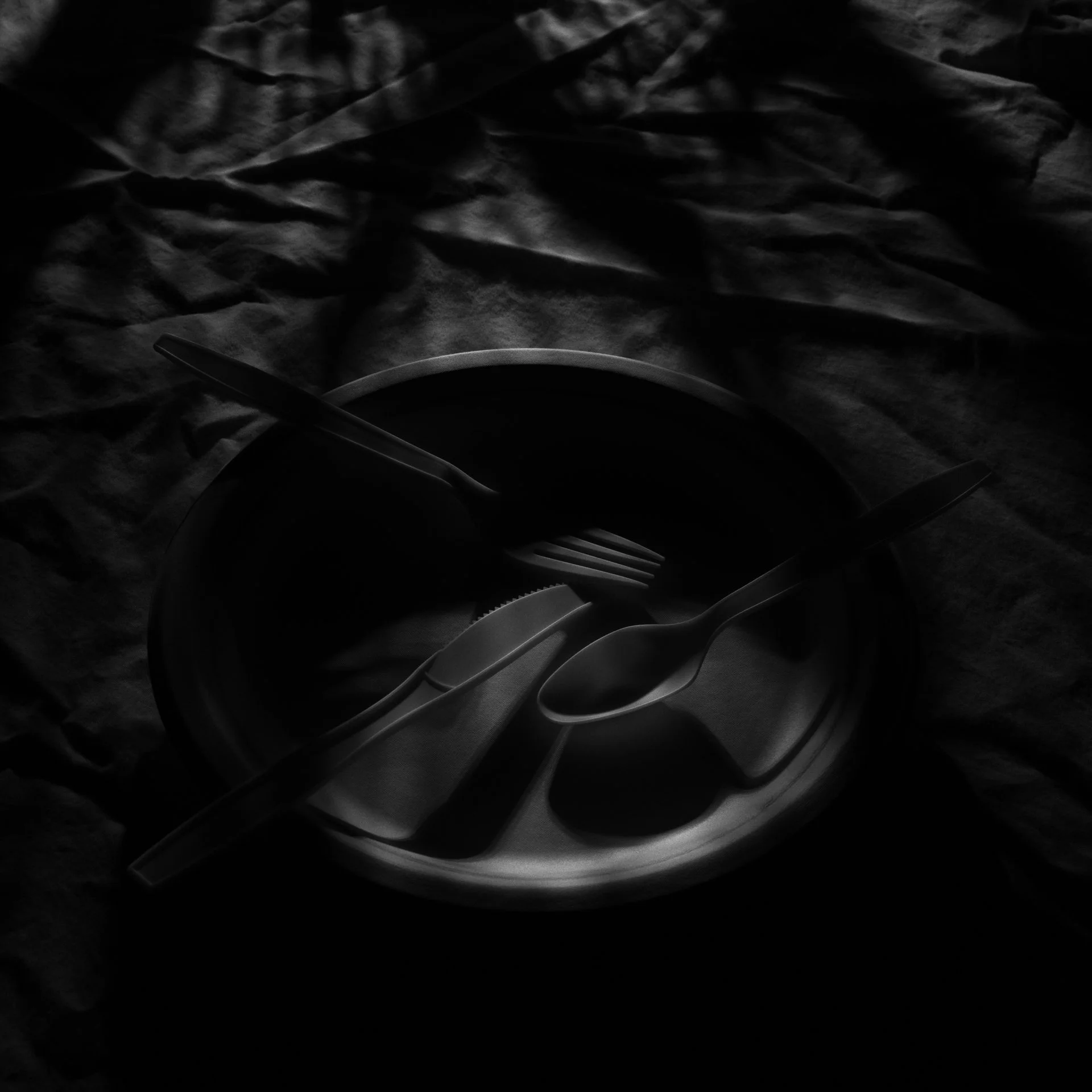  A dark, moody photo shows a paper plate with a plastic fork, knife, and spoon arranged in it, set on a wrinkled fabric surface. Soft light and shadows create a dramatic, high-contrast effect. 