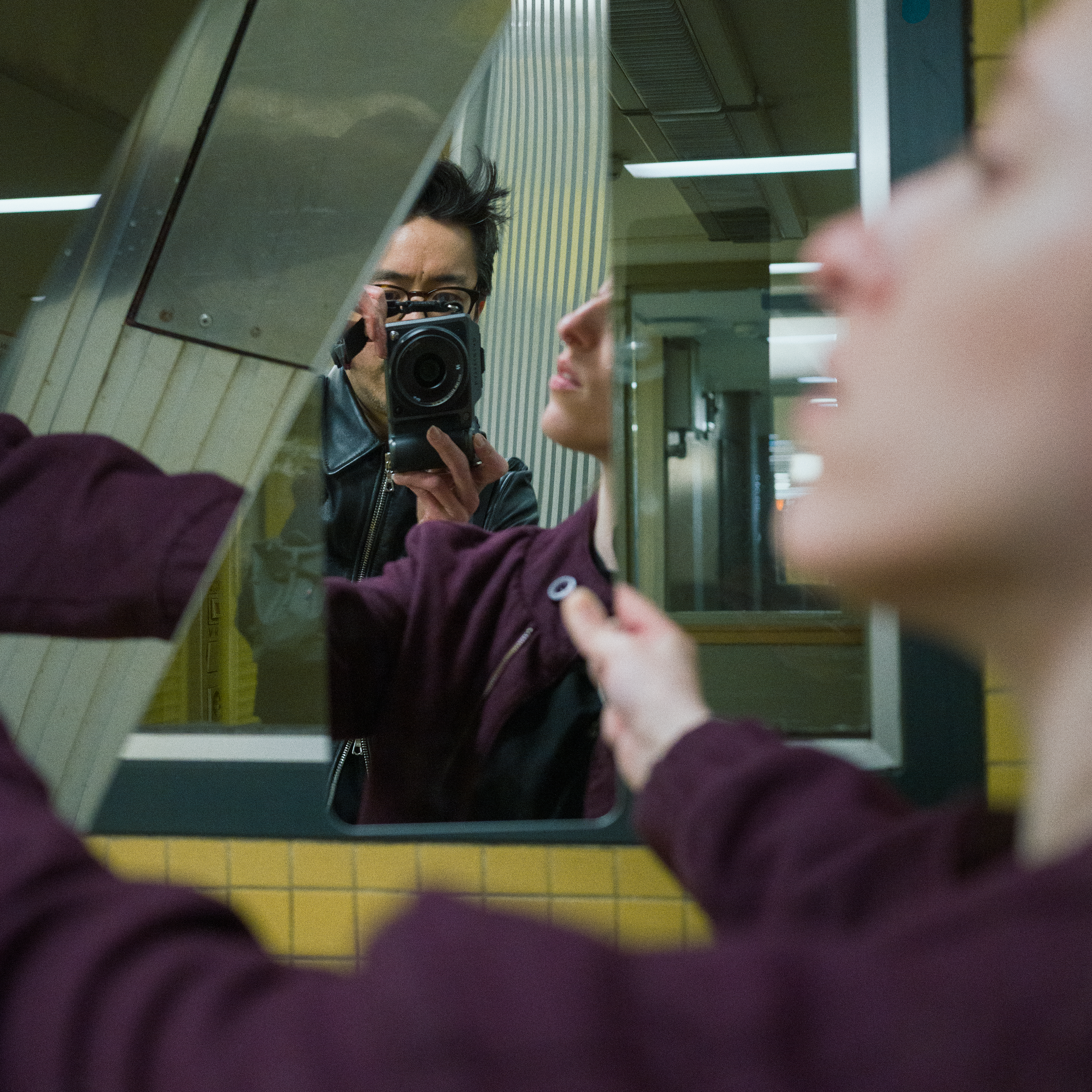 A young woman holds a mirror, reflecting a man taking a photo with a camera. The background features yellow tiled walls and bright indoor lighting of a Berlin subway station. The image captures both reflections and partial profiles.