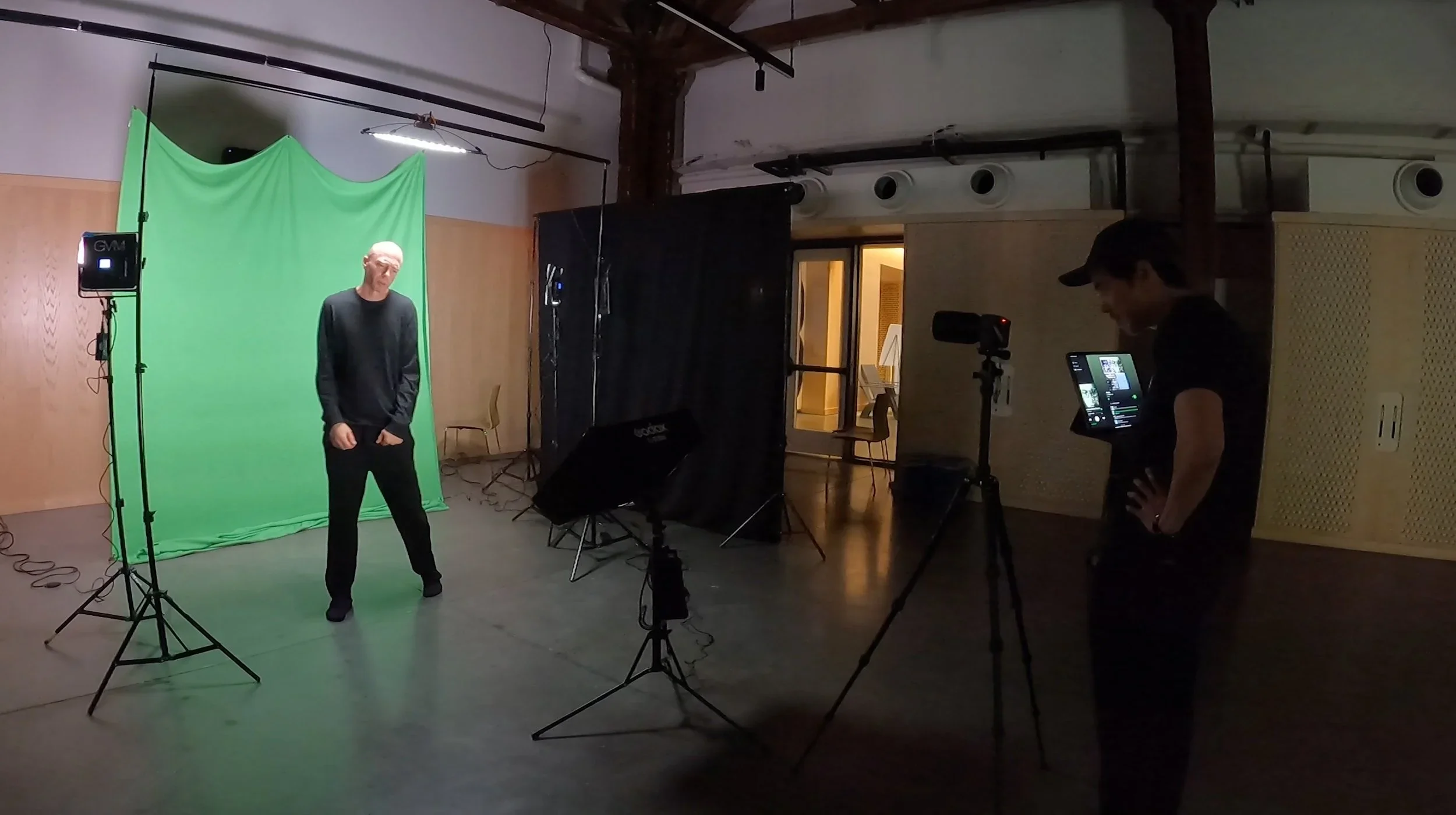 A man stands in front of a green screen in a studio with lighting equipment while another man operates a camera and monitor. The room has exposed beams and an open door to a lit hallway.