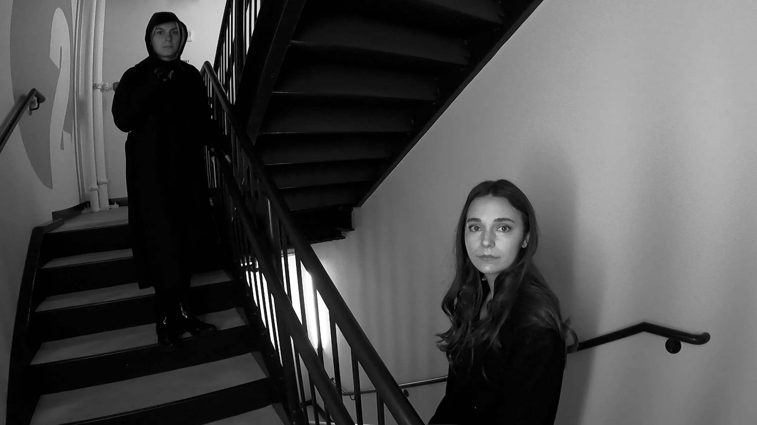 Two young women in dark clothing stand on a staircase indoors; one is at the top step, partially obscured by shadows, while the other stands on the lower steps, looking directly at the camera in a black-and-white setting.
