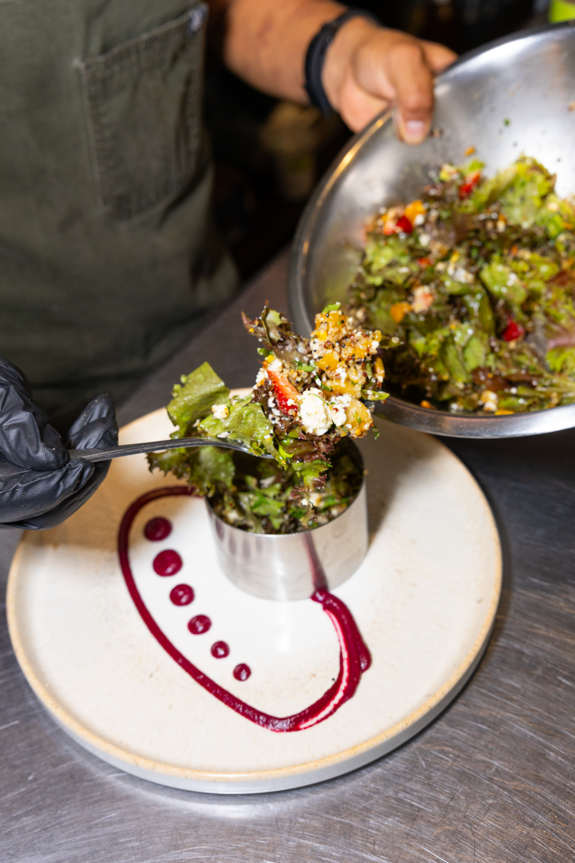 A person in gloves is serving a salad from a stainless steel bowl onto a plate. The salad contains leafy greens, vegetables, and cheese. The plate has red sauce decorative dots and a red curved line.