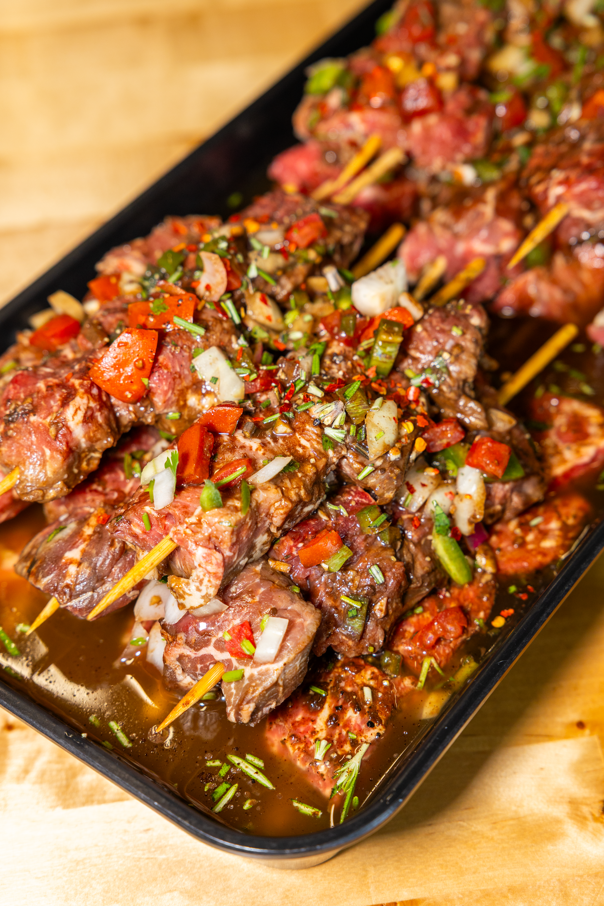 Close-up of marinated beef skewers with chopped vegetables and herbs on black tray, sitting on a wooden surface.