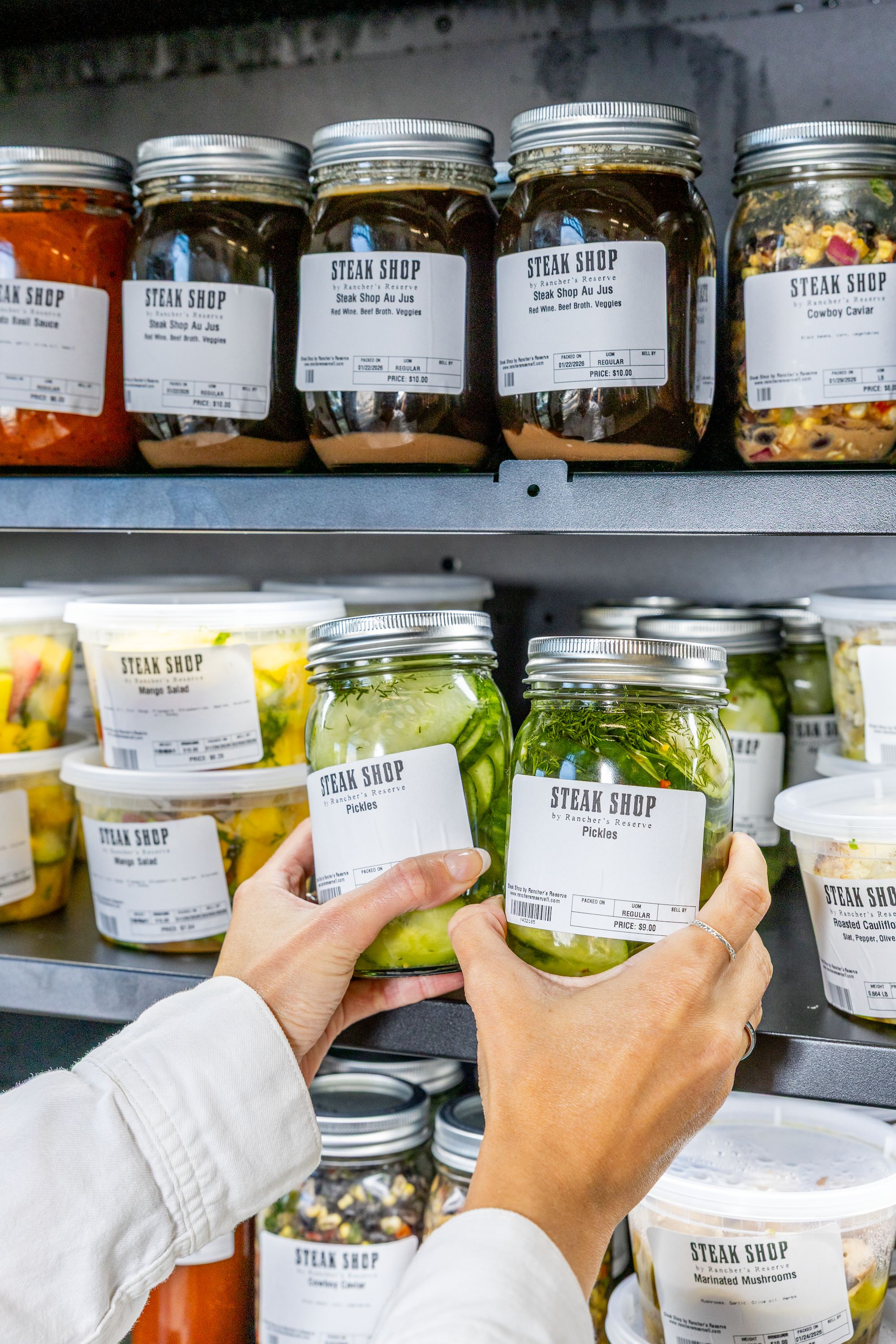 People shopping for jars of pickled vegetables in a grocery store's refrigerated section.