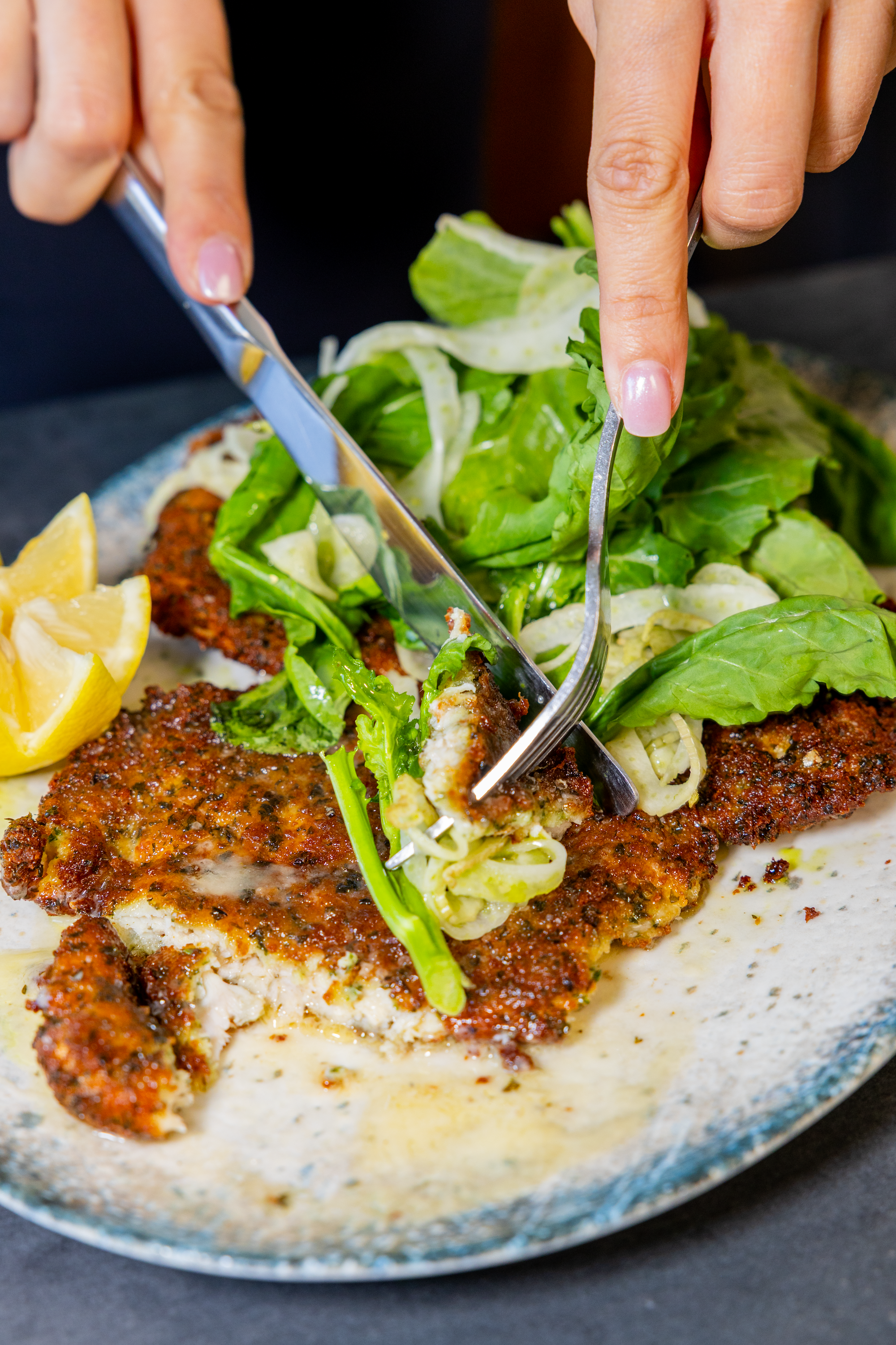 Close-up of a person slicing cooked fish topped with herbs and spices, served with fresh lettuce, lemon wedges, and a creamy sauce in a rustic ceramic dish.