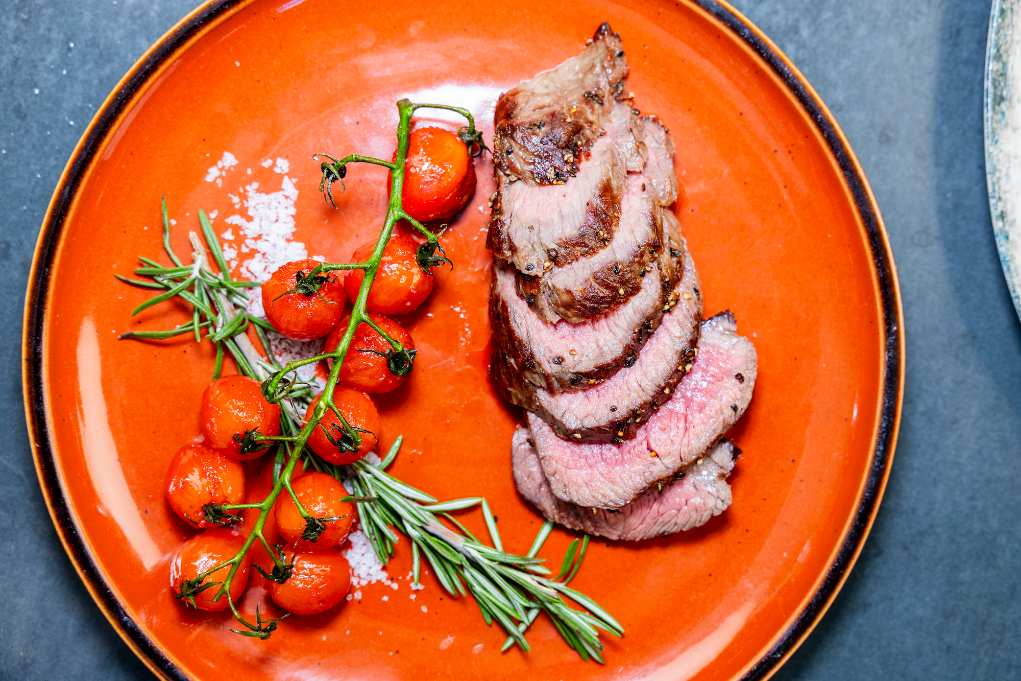 A plate of sliced steak with roasted tomatos on the vine and rosemary and salt on a red plate