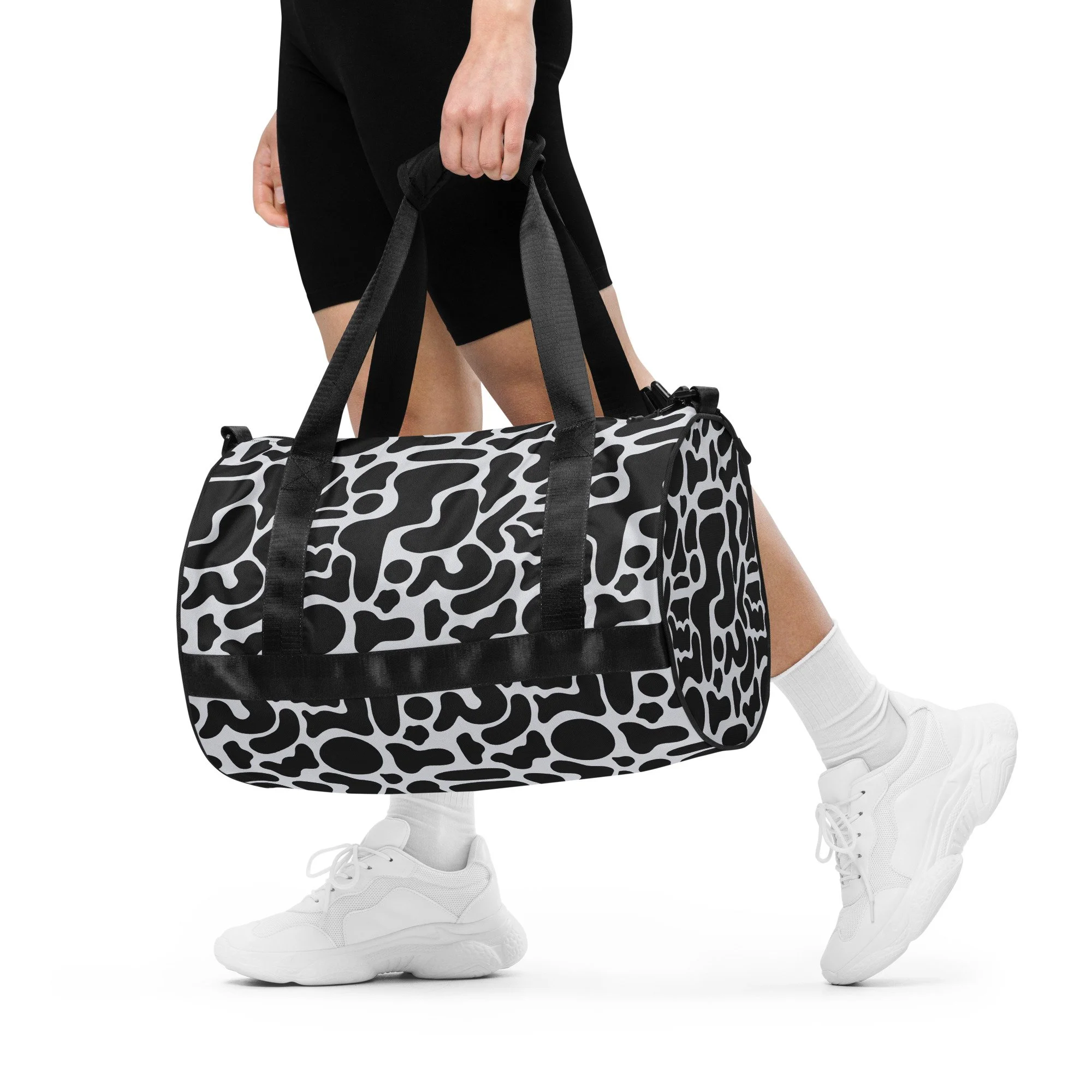 Person carrying a black and white patterned duffel bag, wearing black shorts and white sneakers.