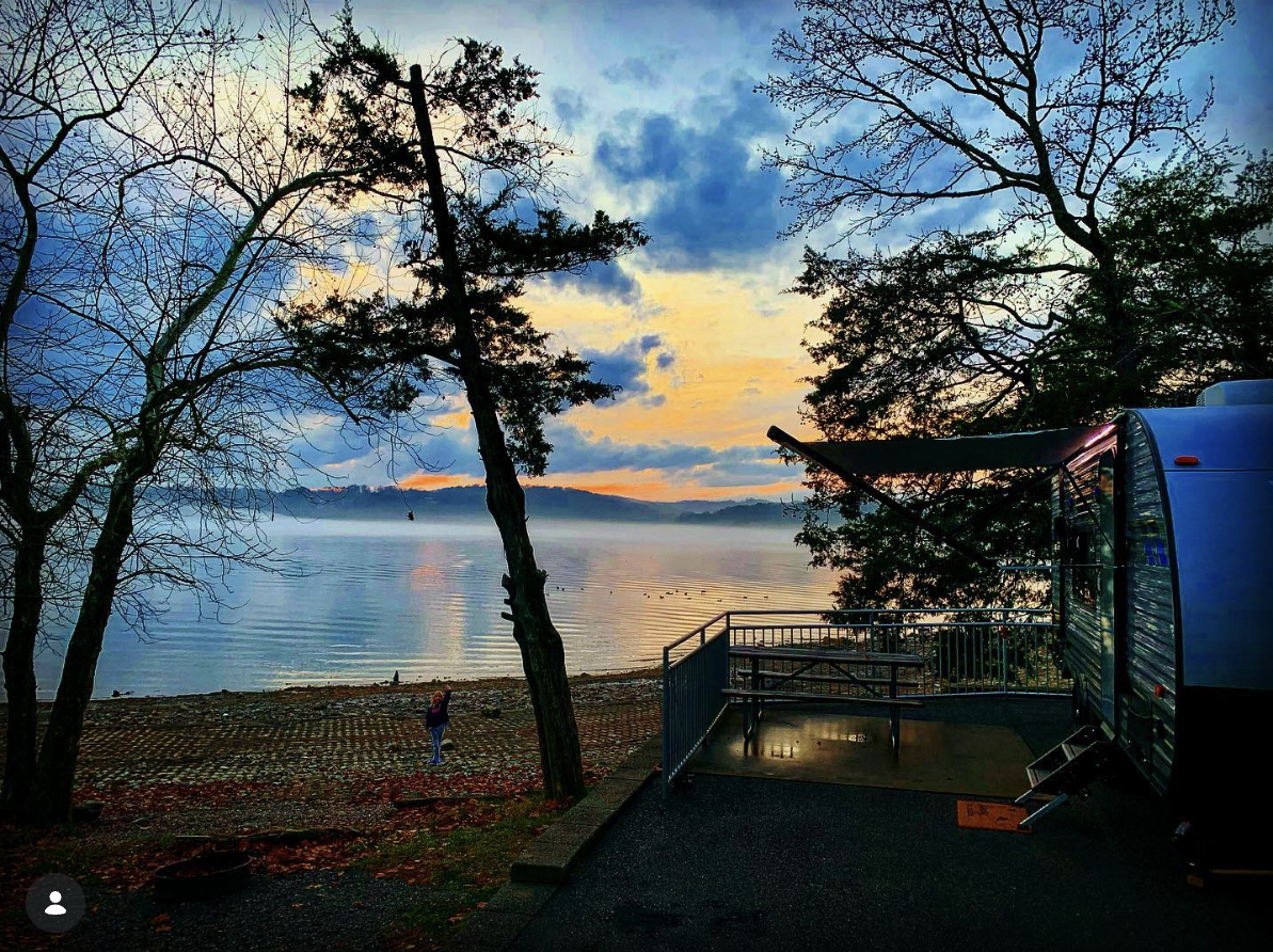 A lakeside scene at sunset with trees, a person taking a photo, and an RV with an extended awning on the right.