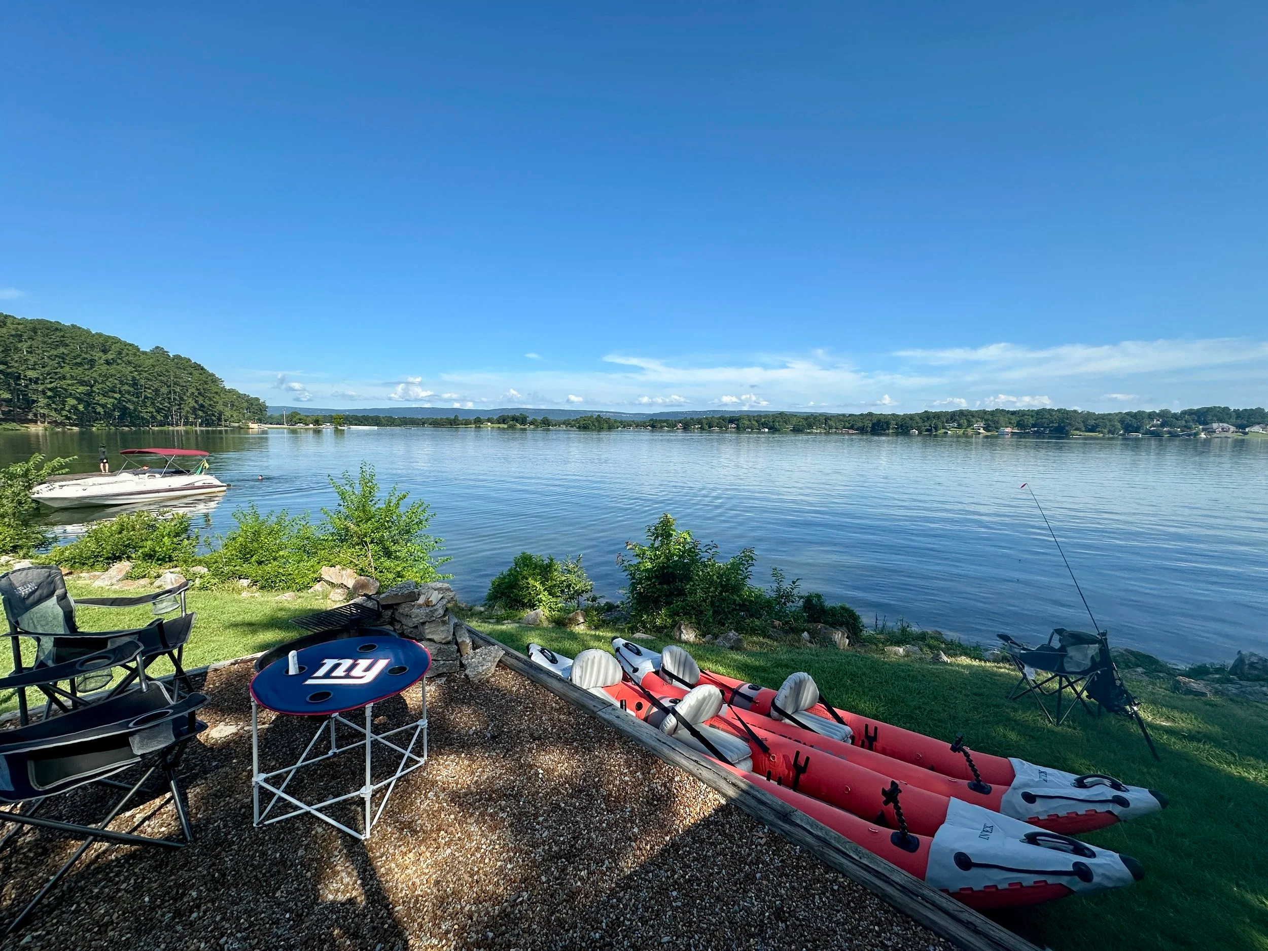 A lakeside scene with boats, kayaks, and outdoor chairs on a grassy area, overlooking a calm lake under a partly cloudy sky with a wooded shoreline in the background.