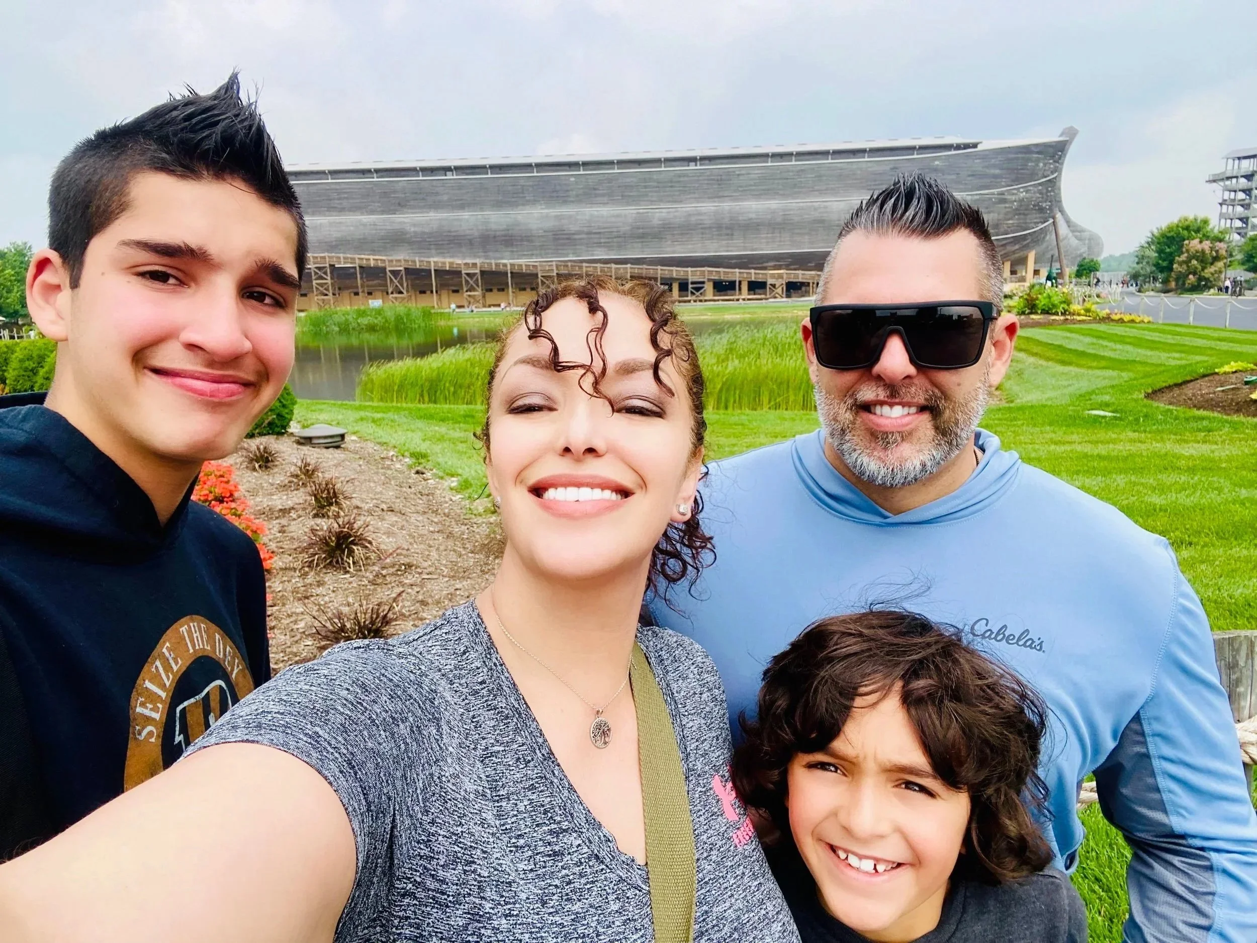 Group of four smiling people taking a selfie outdoors with a large building in the background, greenery, and a pond.