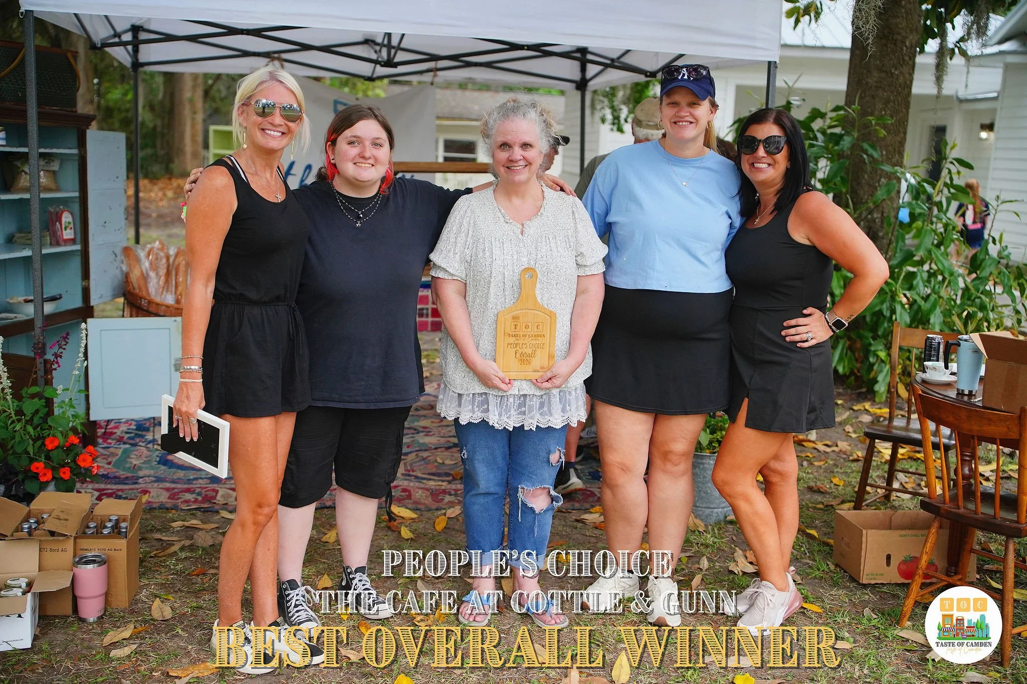 Group of five women standing outdoors under a canopy, celebrating the 'Best Overall Winner' at Cottle & Gunn, with one woman holding a wooden plaque. There are boxes and tables with items around them.