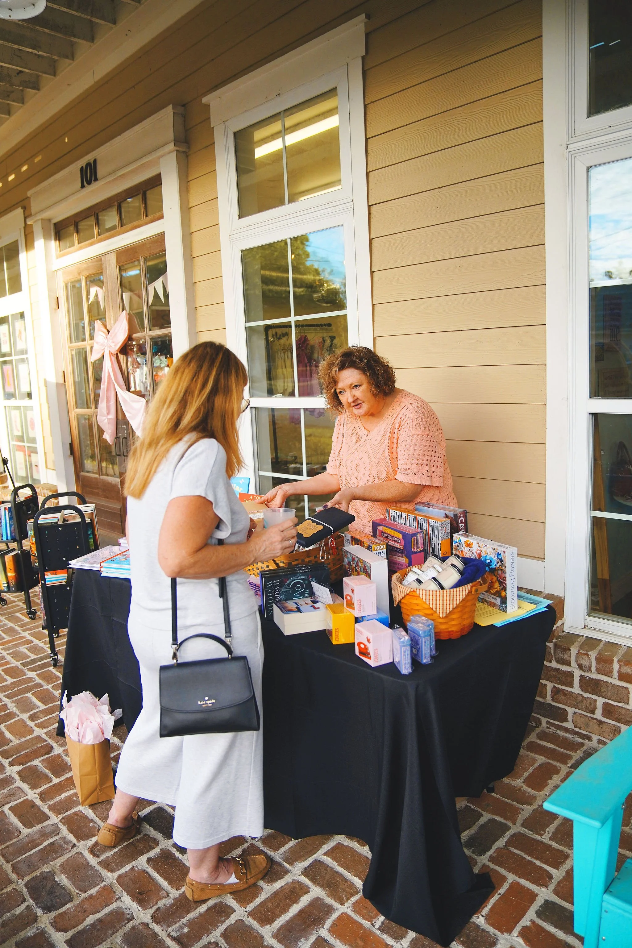 Woman shopping at an outdoor sale table with small items and books, talking to a vendor in front of a beige building with large windows and a brick floor.