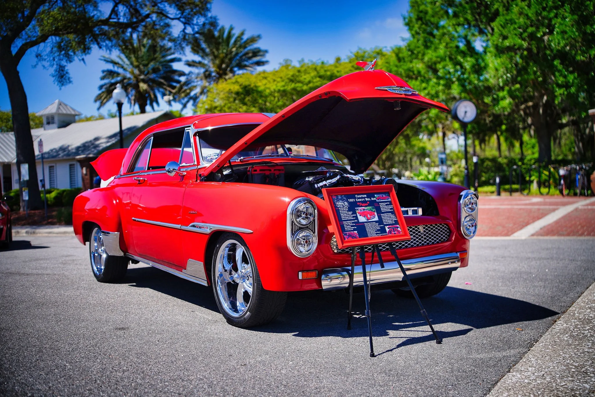 Red vintage Chevrolet car with open hood at an outdoor car show, display board in front, trees and buildings in background on a sunny day.