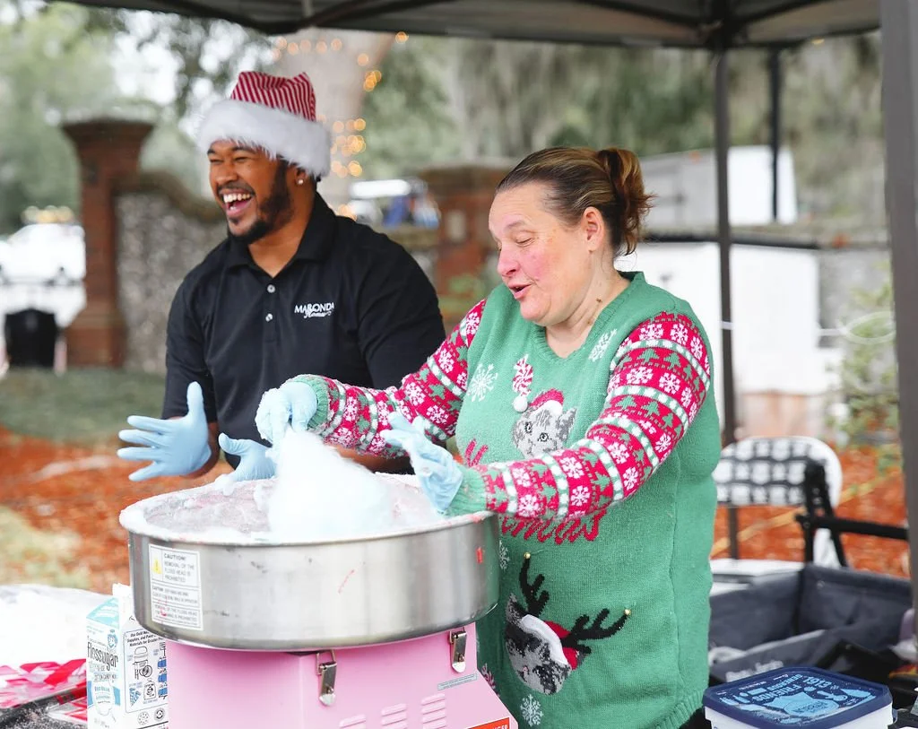 A man wearing a Santa hat and a woman with a holiday sweater making cotton candy outdoors.
