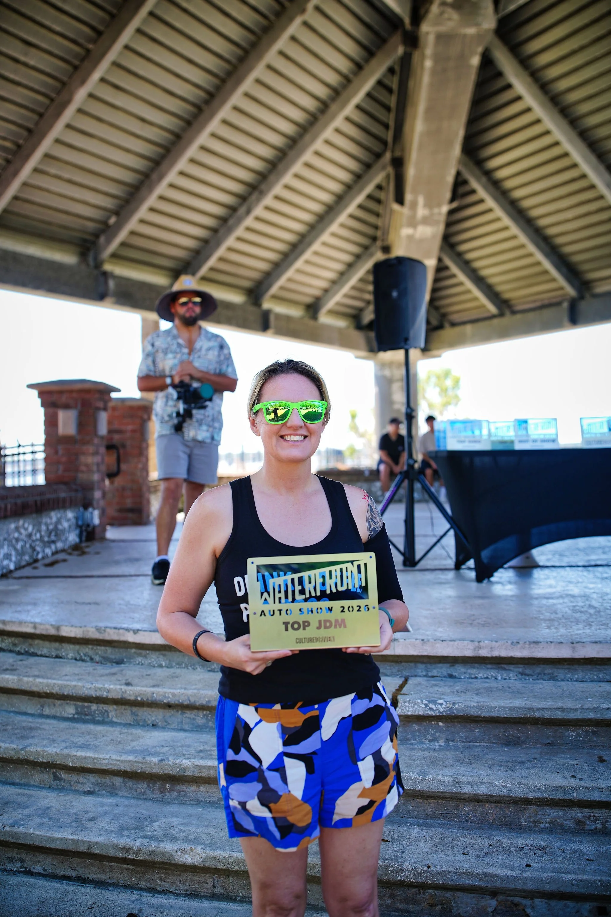 A woman wearing sunglasses and a black tank top holding a sign that reads "WaterRun! Auto Show 2026" with "Top JDM" at the bottom, smiling on a stage with another person in the background under a pavilion.