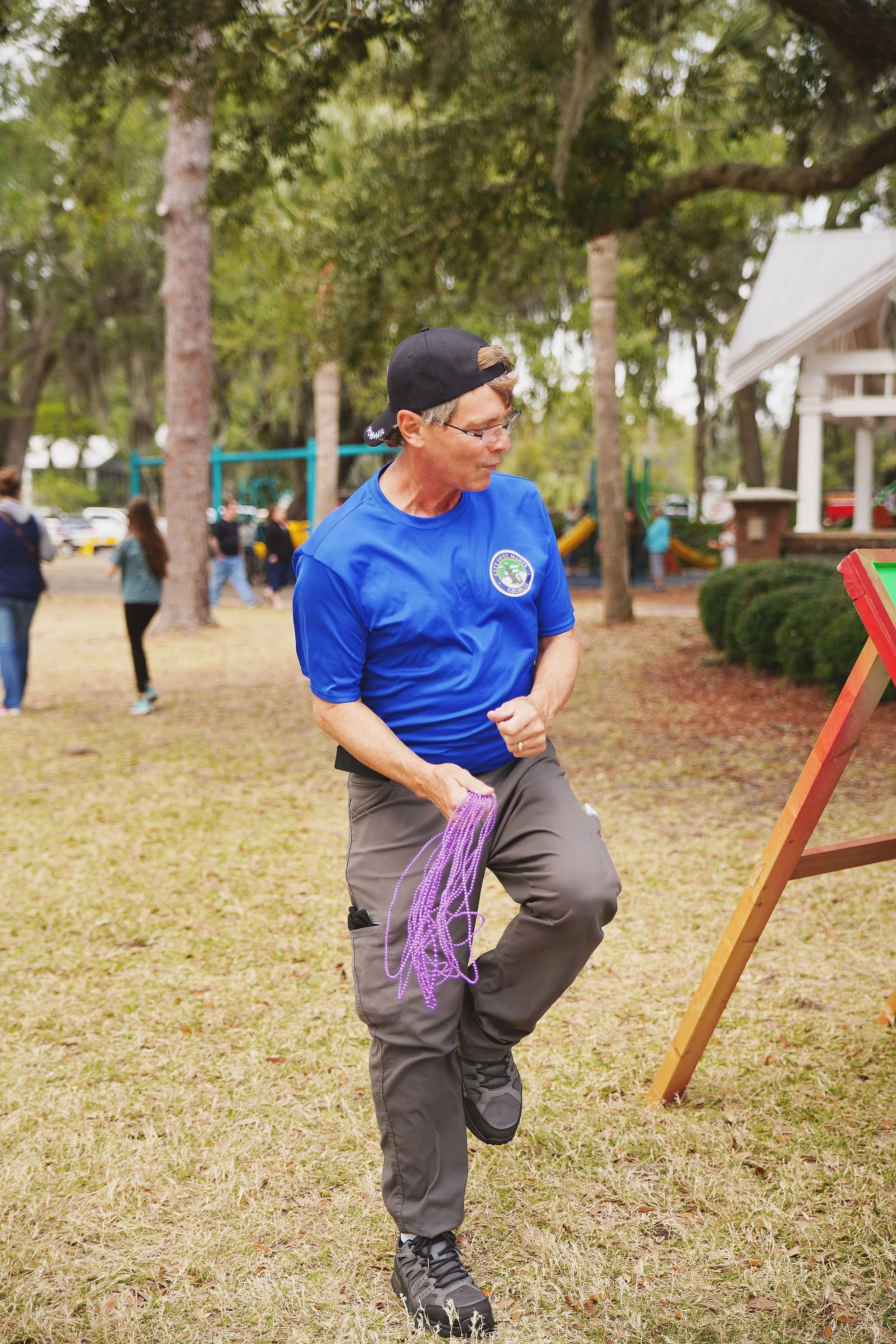 A man in a blue shirt and gray pants holding purple beads, standing outdoors on a grassy area.