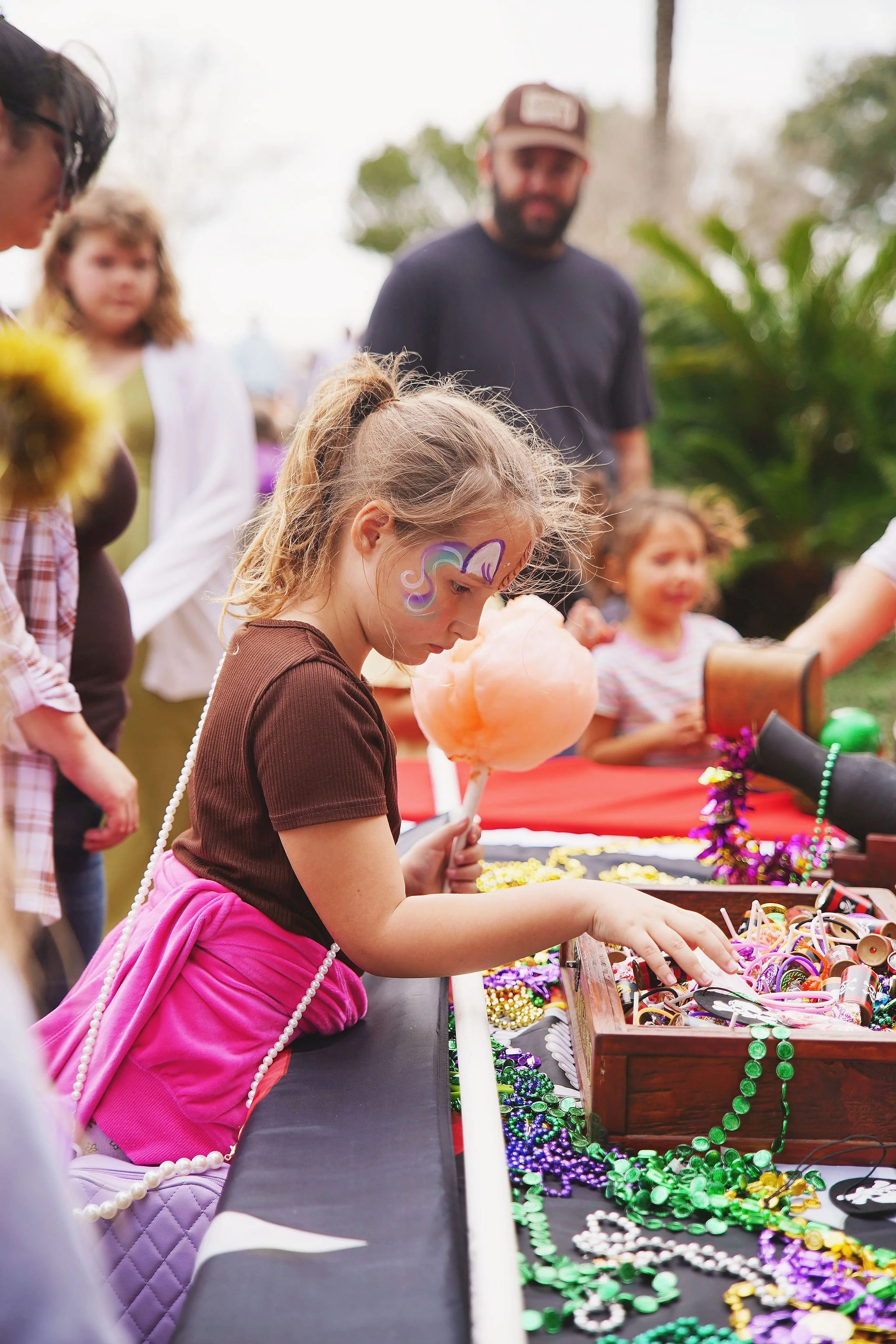 A young girl with a unicorn painted on her face is at a booth with Mardi Gras beads, jewelry, and accessories at an outdoor event, surrounded by other children and adults.