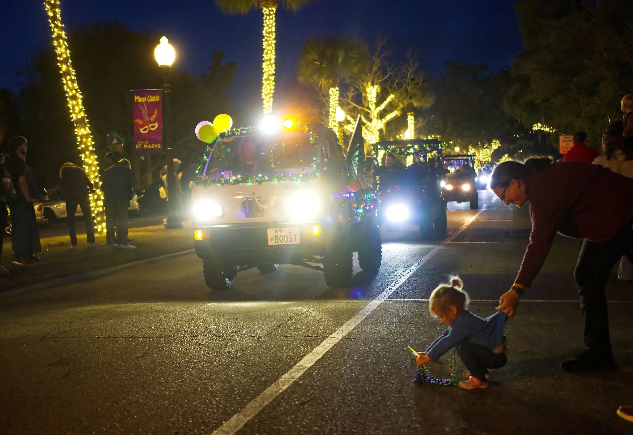 Nighttime parade with decorated vehicles, illuminated trees, and people watching along the street. A young child and an adult are near the curb, with the child looking at a phone, while the adult holds the child's hand. A festive atmosphere with ligh