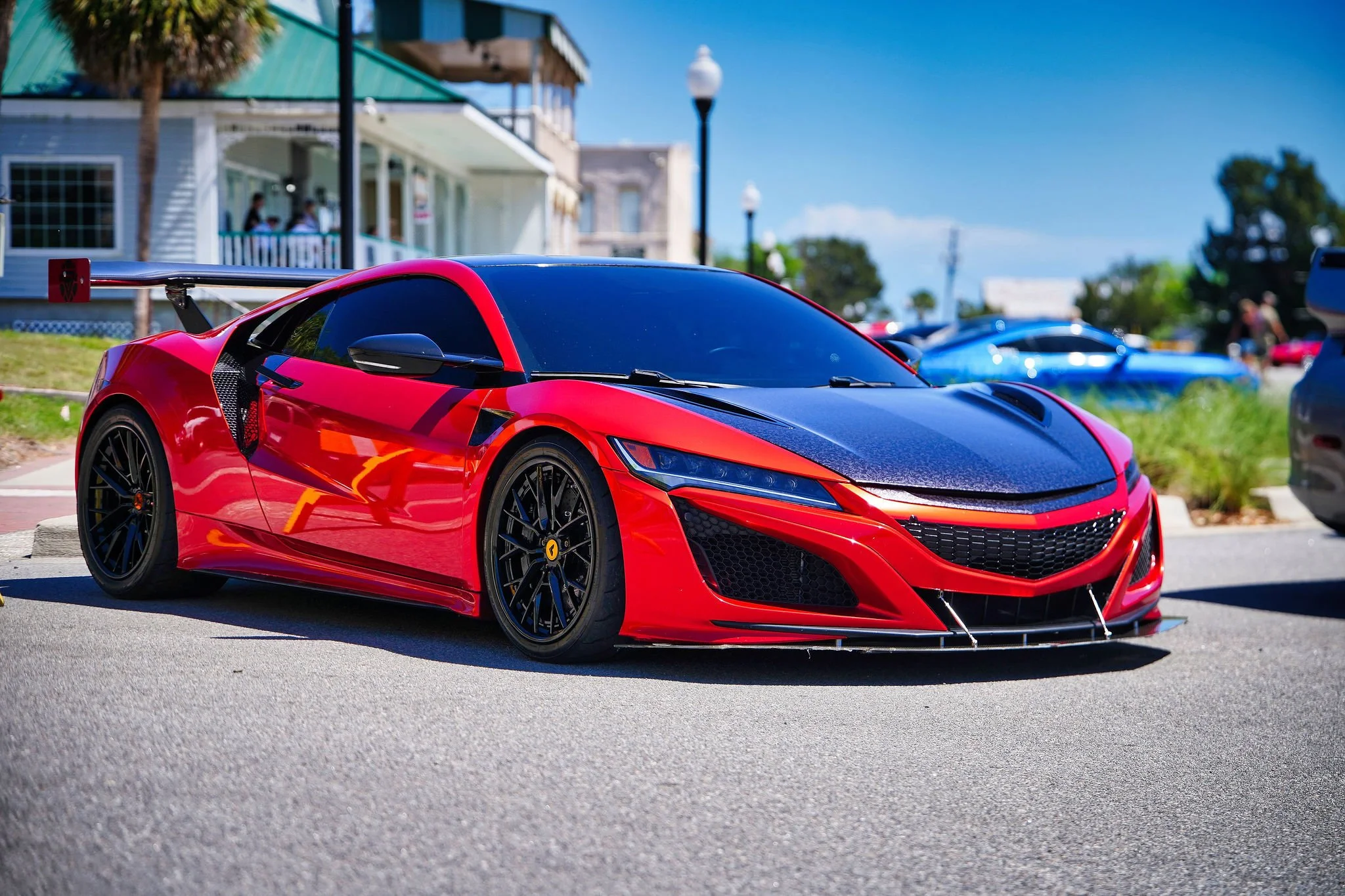 A red sports car with black accents parked on a street during daytime. In the background, there are other cars, a building with a balcony, and a street lamp under a clear blue sky.