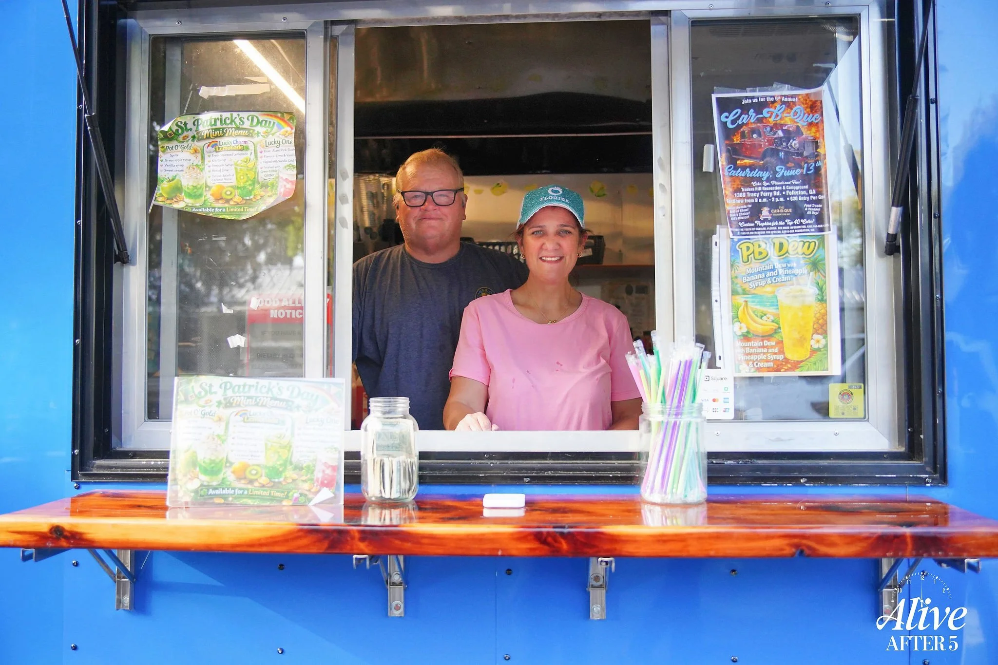 Two people smiling at a food truck window. The woman in front is wearing a pink shirt and a cap, and the man behind her is wearing glasses and a gray shirt. The food truck has posters advertising St. Patrick's Day mini menu and beverages.