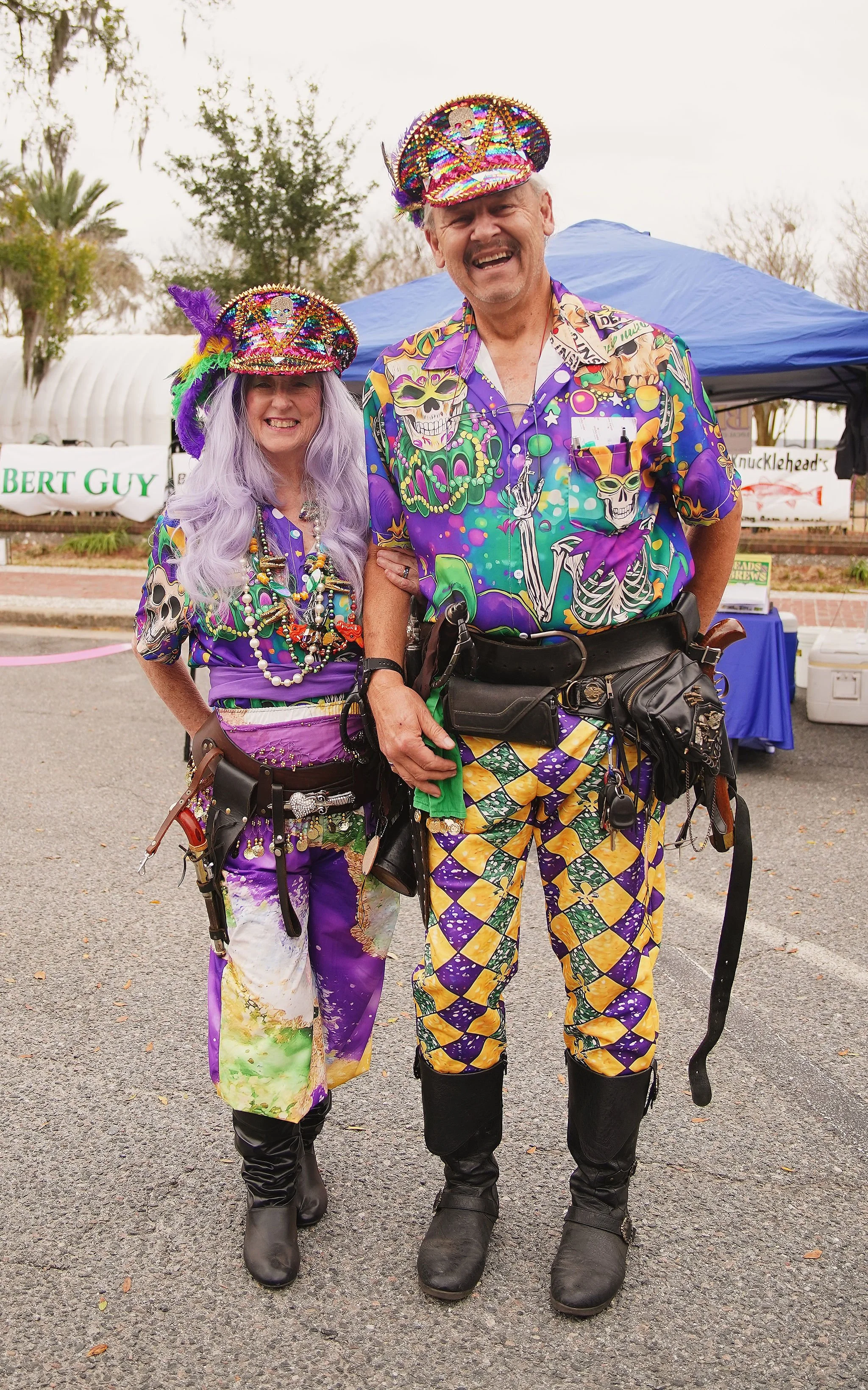 Two people dressed in colorful, festive costumes with skeleton and skull motifs, wearing decorated hats, standing outdoors at a festival or event.