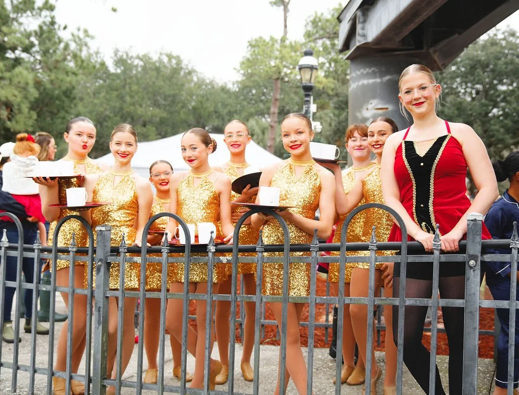 A group of young female performers in gold and red costumes standing behind a black fence, holding white cups on trays and smiling.