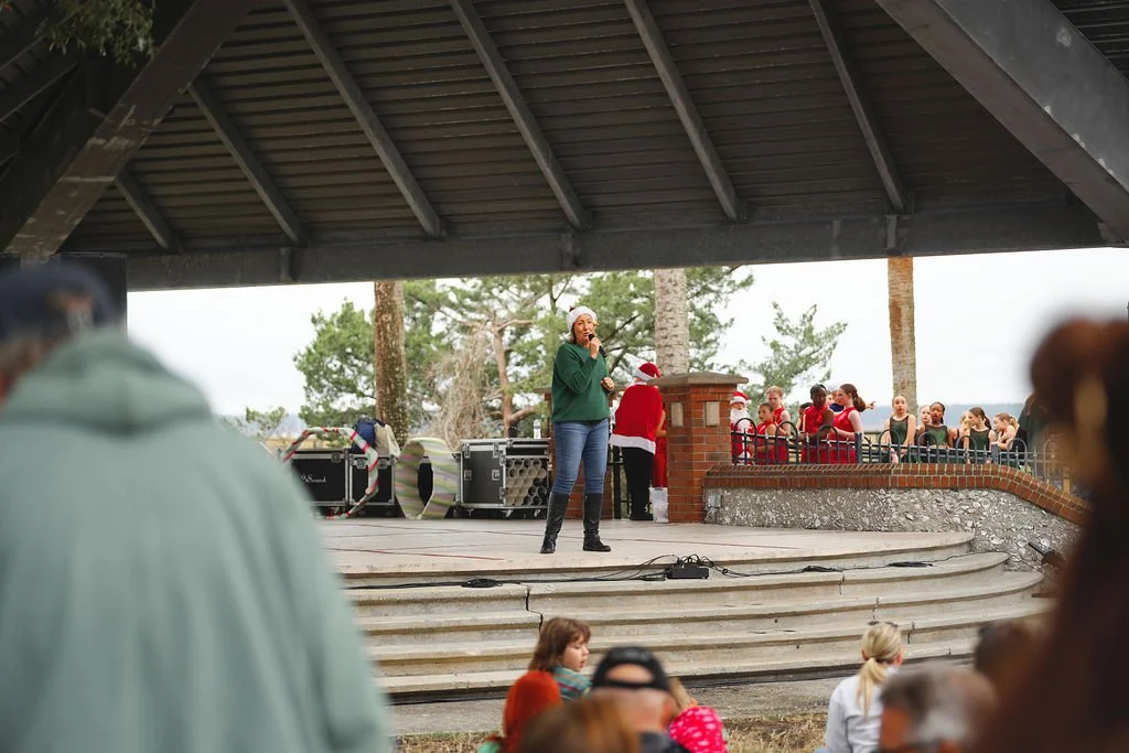 Outdoor Christmas event with a woman singing on stage, shouted by a person dressed as Santa Claus, with children and adults watching nearby.