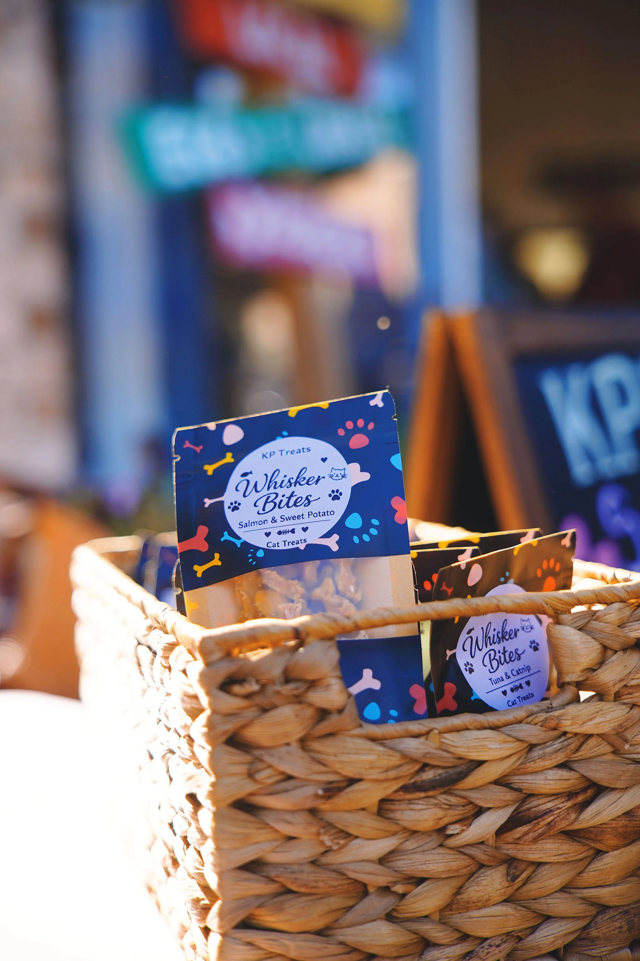 Close-up of a woven basket filled with cat treats labeled "Whisker Bites" in two flavors, salmon and sweet potato, and tuna and catnip, set outdoors with colorful signs and blackboards in the blurred background.