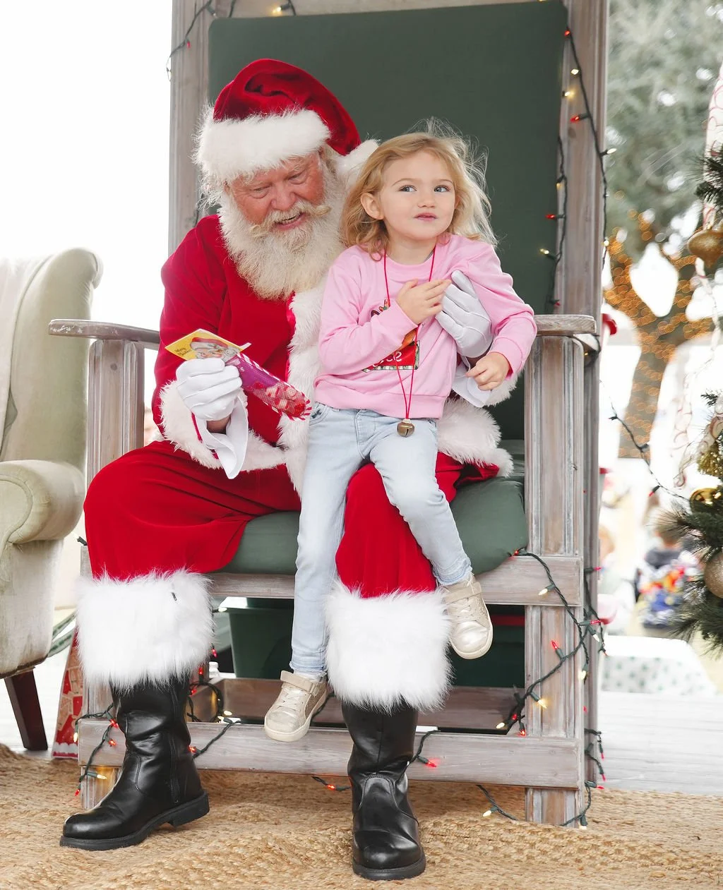 A young girl sitting on Santa Claus's lap at a Christmas event. Santa is wearing a red suit with white fur trim, black boots, and a Santa hat. The girl is wearing a pink long sleeve shirt and light blue pants. They are inside a decorated setting with
