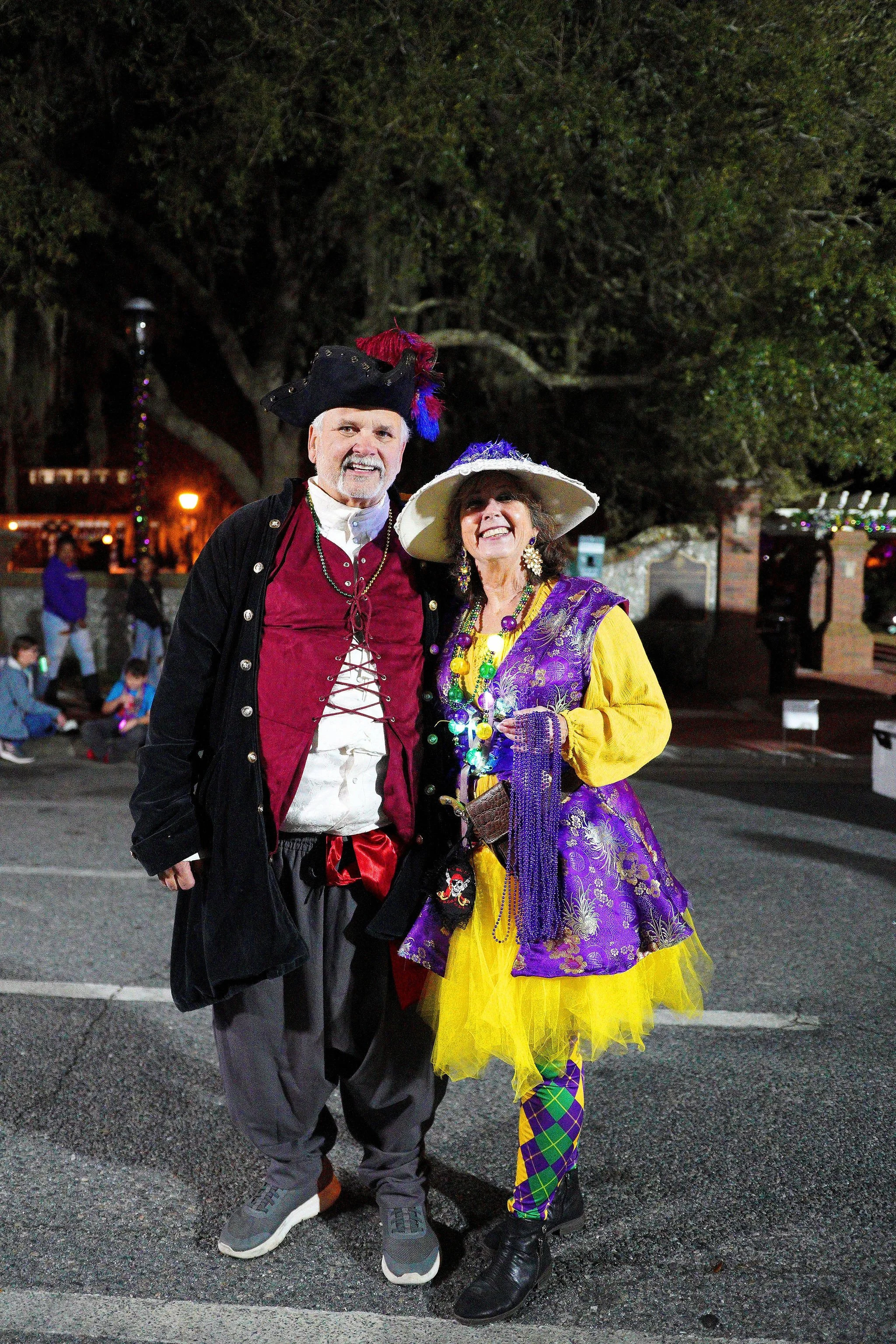 Two people dressed as pirates and whimsical characters, smiling outdoors at night with trees and other people in the background.
