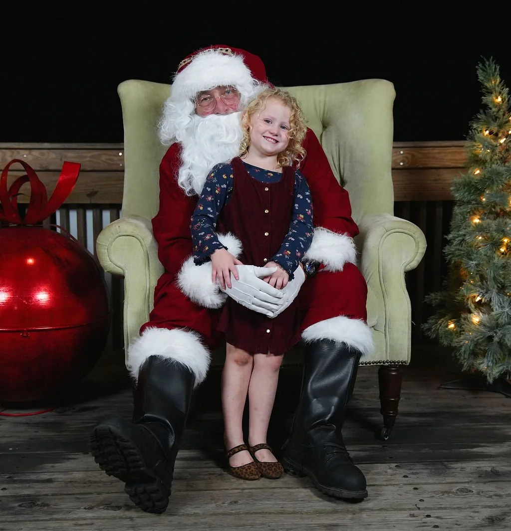A young girl with curly blonde hair sitting on Santa Claus's lap, both smiling, in a festive setting with Christmas decorations, a large red ornament, and a Christmas tree with lights.