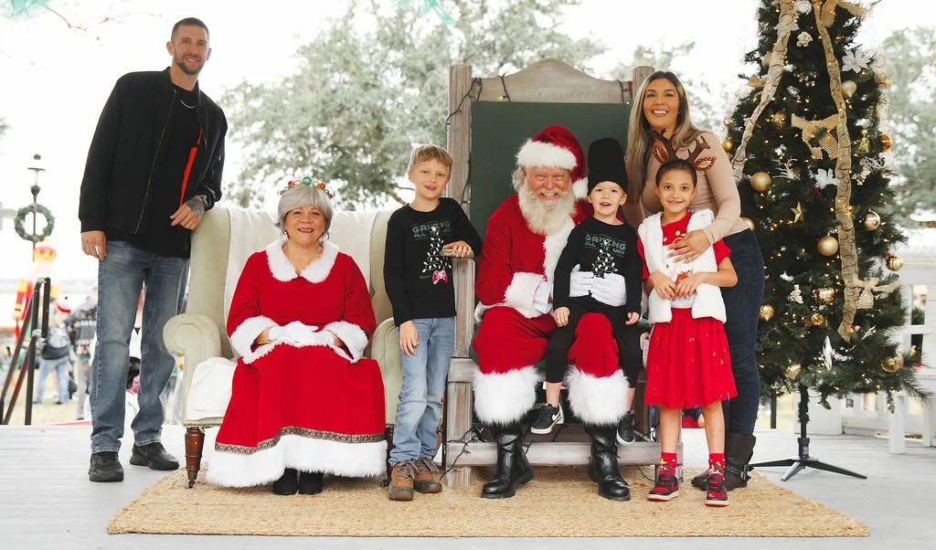 Family posing with Santa Claus and Mrs. Claus outdoors during Christmas, with a decorated Christmas tree nearby.
