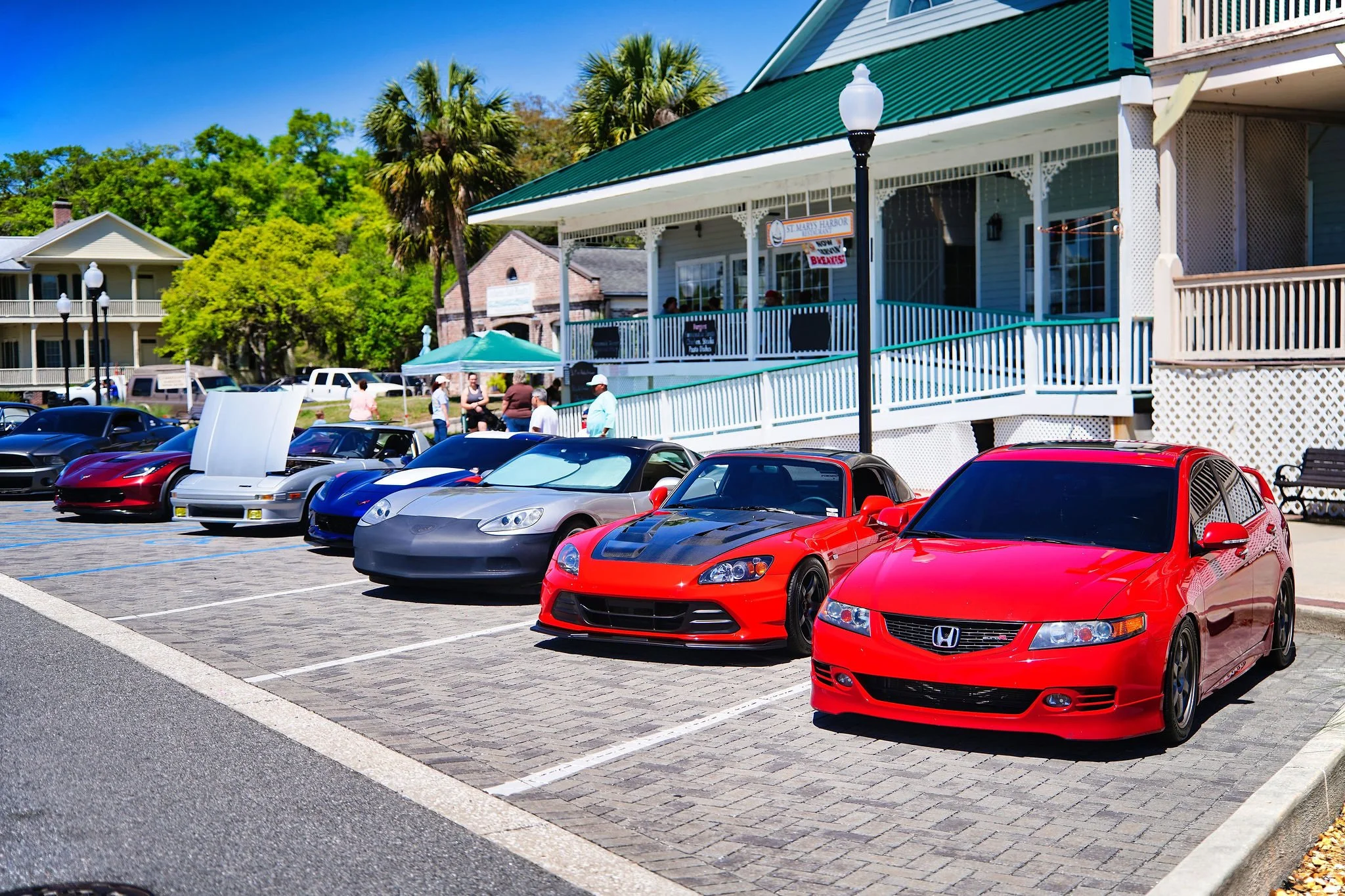 Lineup of luxury and sports cars parked in front of a restaurant with outdoor seating and people walking nearby. The cars include a red Honda, a gray car, and several others including a Corvette and Tesla, with a sunny day and trees in the background