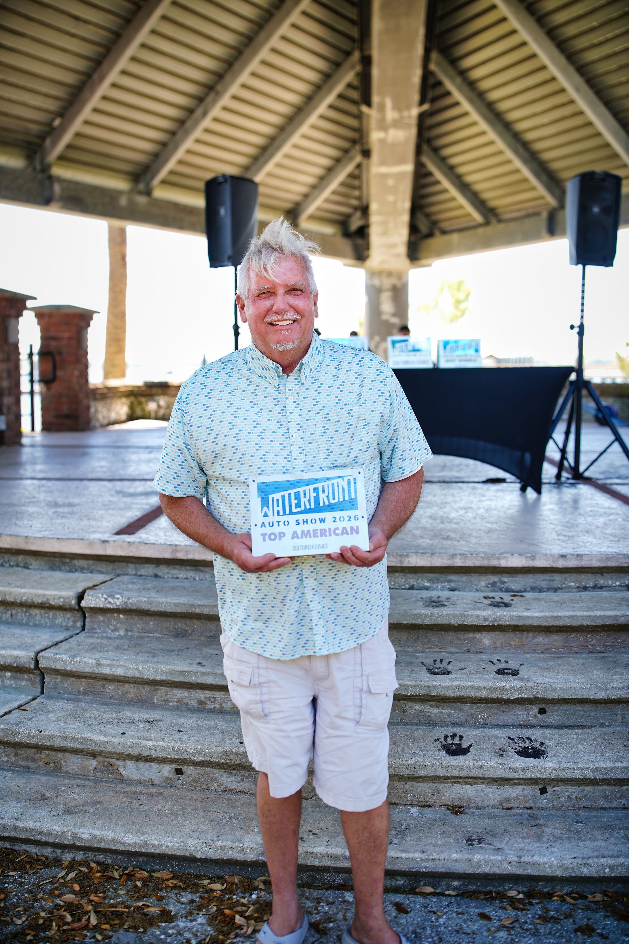 A man standing on outdoor steps holding an award plaque, wearing a light blue patterned shirt and beige shorts, smiling with a stage and speakers in the background.