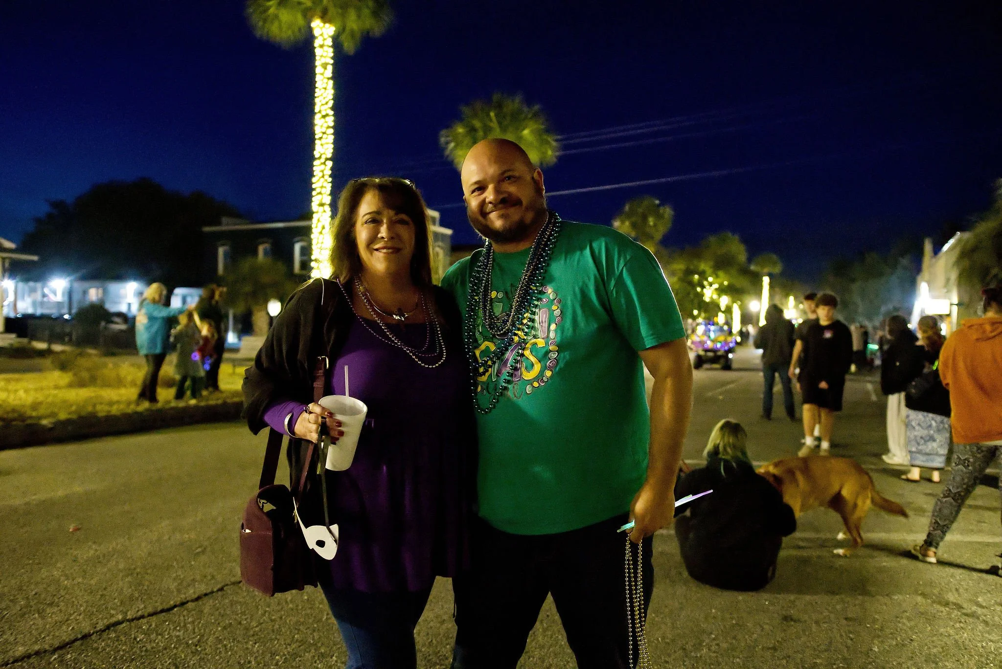 A woman and a man with Mardi Gras beads smiling at night during a festival, with other people, a dog, and illuminated buildings in the background.