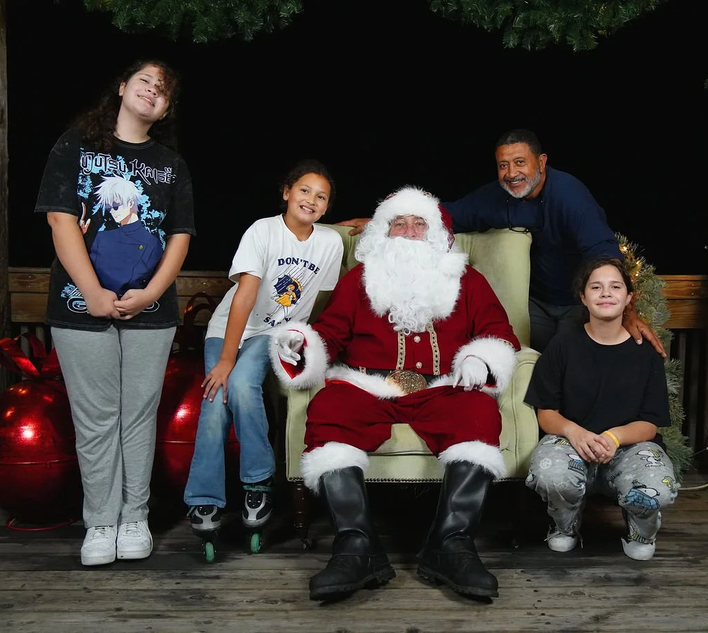 Children sitting with Santa Claus, who is in a green chair, on a wooden deck decorated for Christmas, with a man and a girl standing beside them.