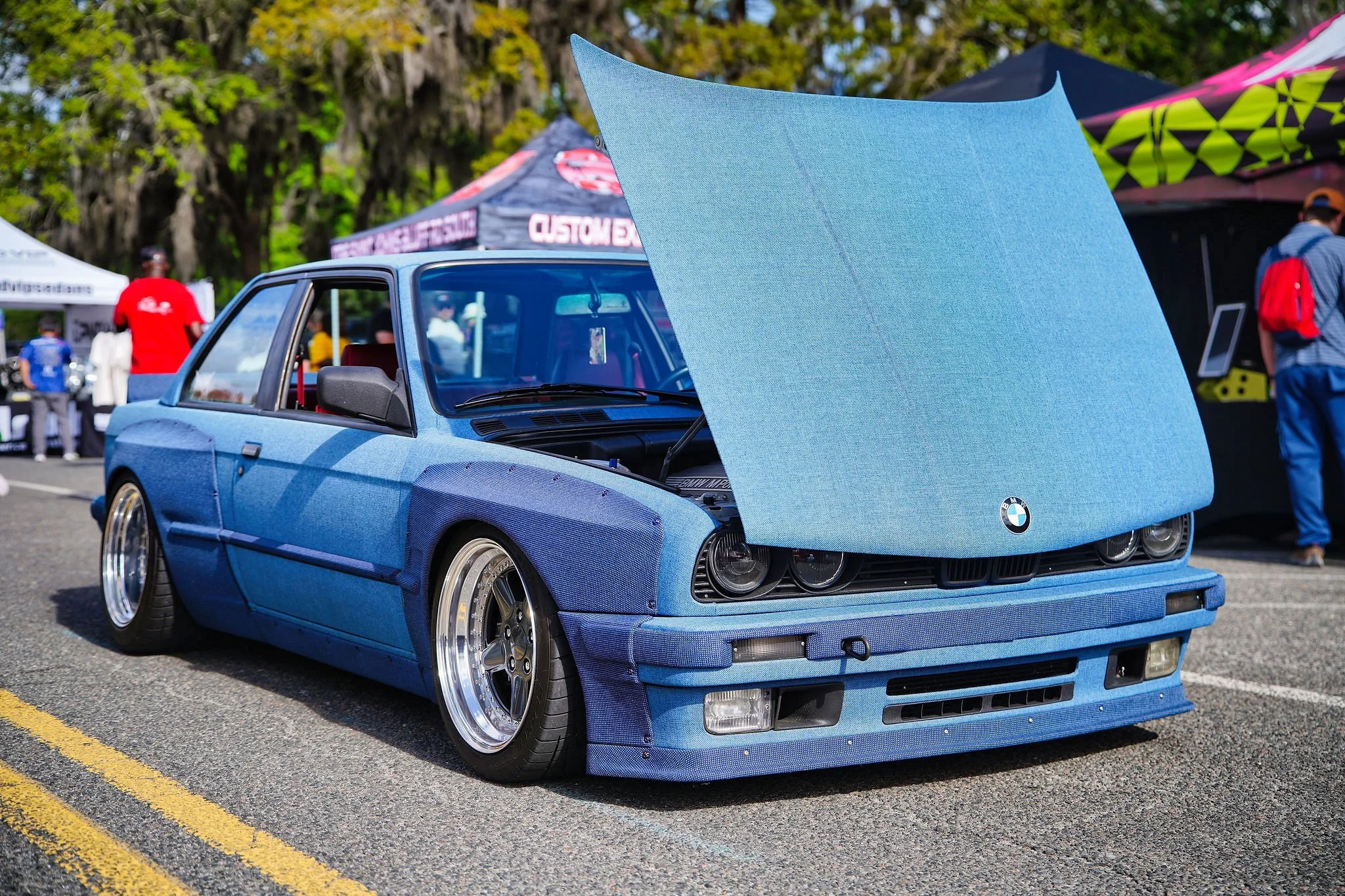 A custom BMW car with a denim fabric exterior at a car show, with its hood open, surrounded by tents and people.