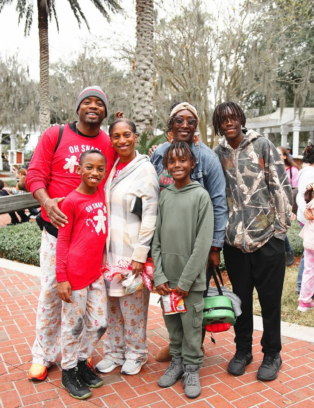 A group of six people, including children and adults, are smiling and standing outdoors in a park with trees and other people in the background.