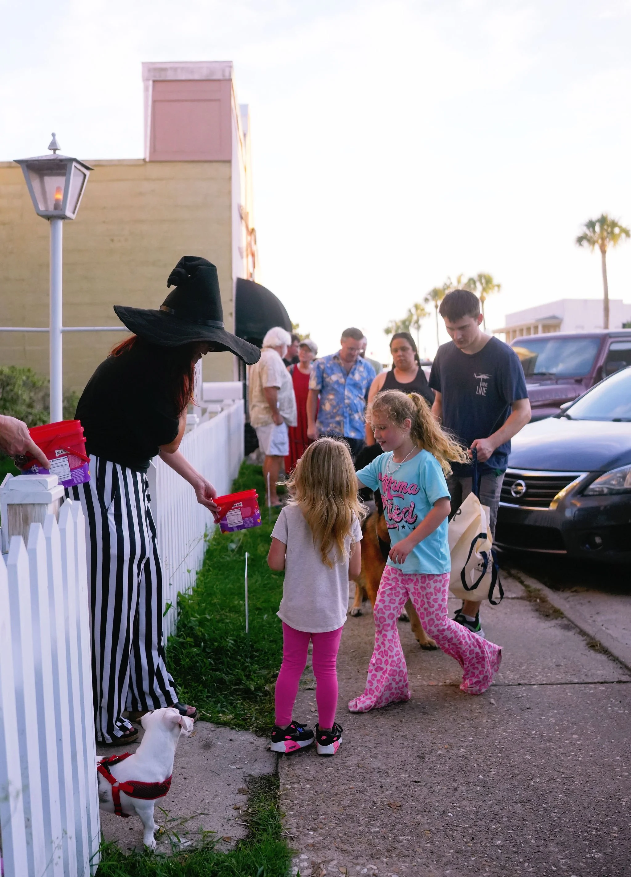 A group of children and adults outdoors near a white picket fence, with a woman dressed as a witch handing out candy, and a small dog wearing a red harness standing on the grass.
