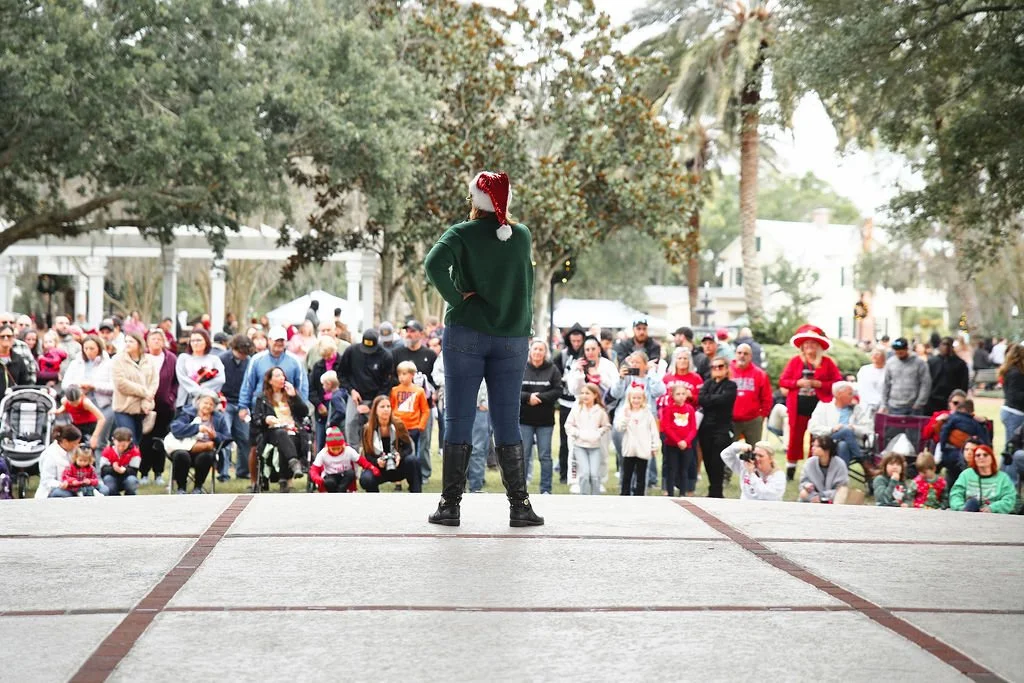 Woman in green sweater and jeans standing on stage with her back facing the audience, wearing a red Santa hat, with a crowd watching in an outdoor park during holiday event.