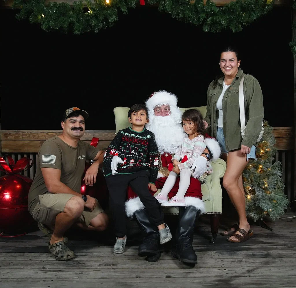 A family with two children sitting on Santa Claus's lap during Christmas, with festive decor including a Christmas tree and ornaments.