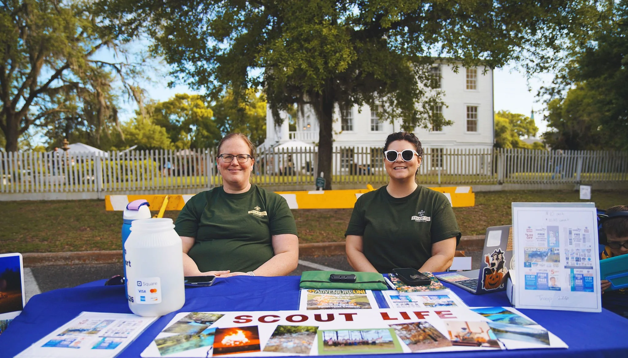 Two women sitting behind a table at a scout event outdoors, with a large white house and trees in the background. The table displays various informational materials, flyers, and signs including a 