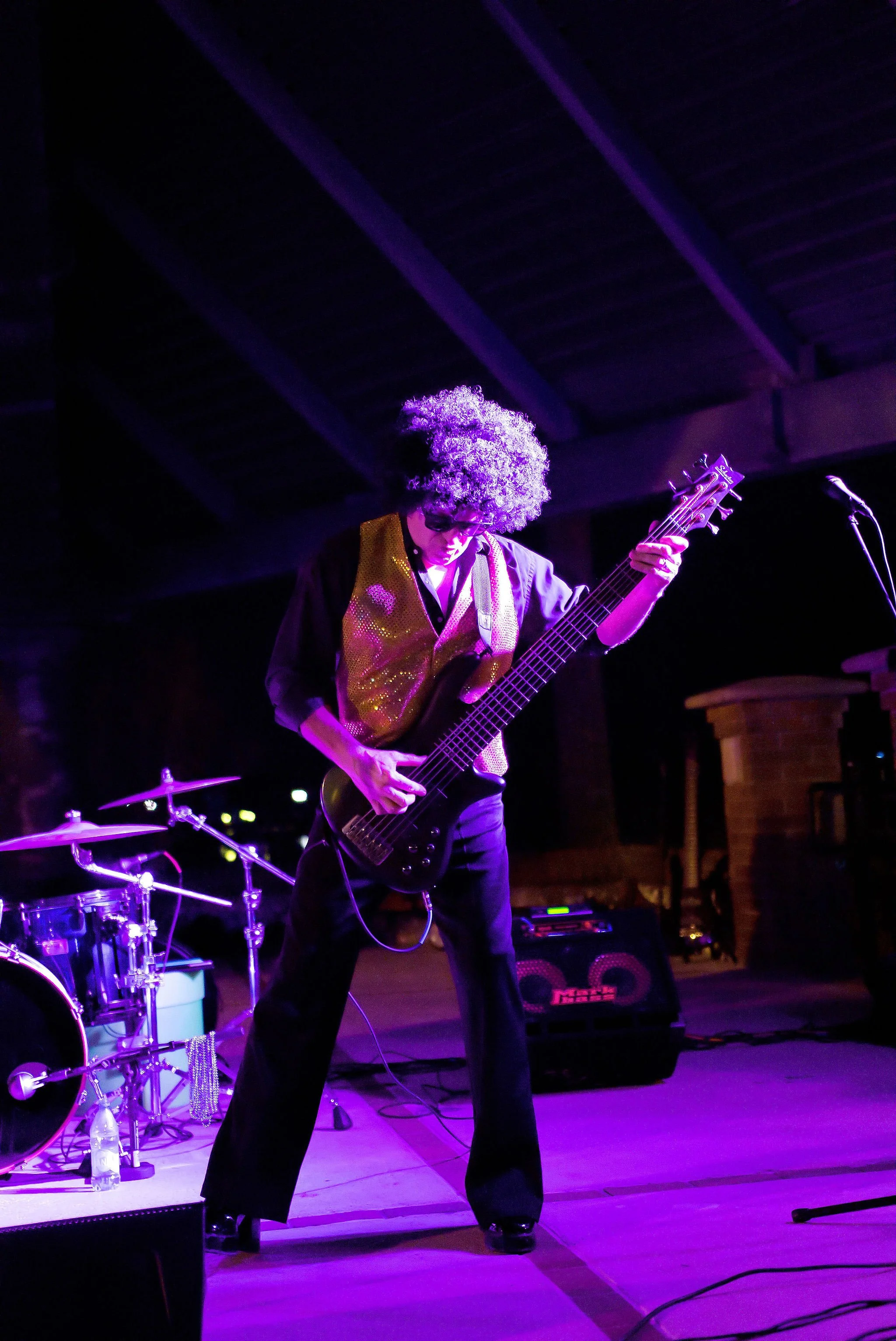 A musician with curly hair and sunglasses playing an electric guitar on stage, dressed in a shiny vest and black pants, with a drum set in the background under purple lighting.