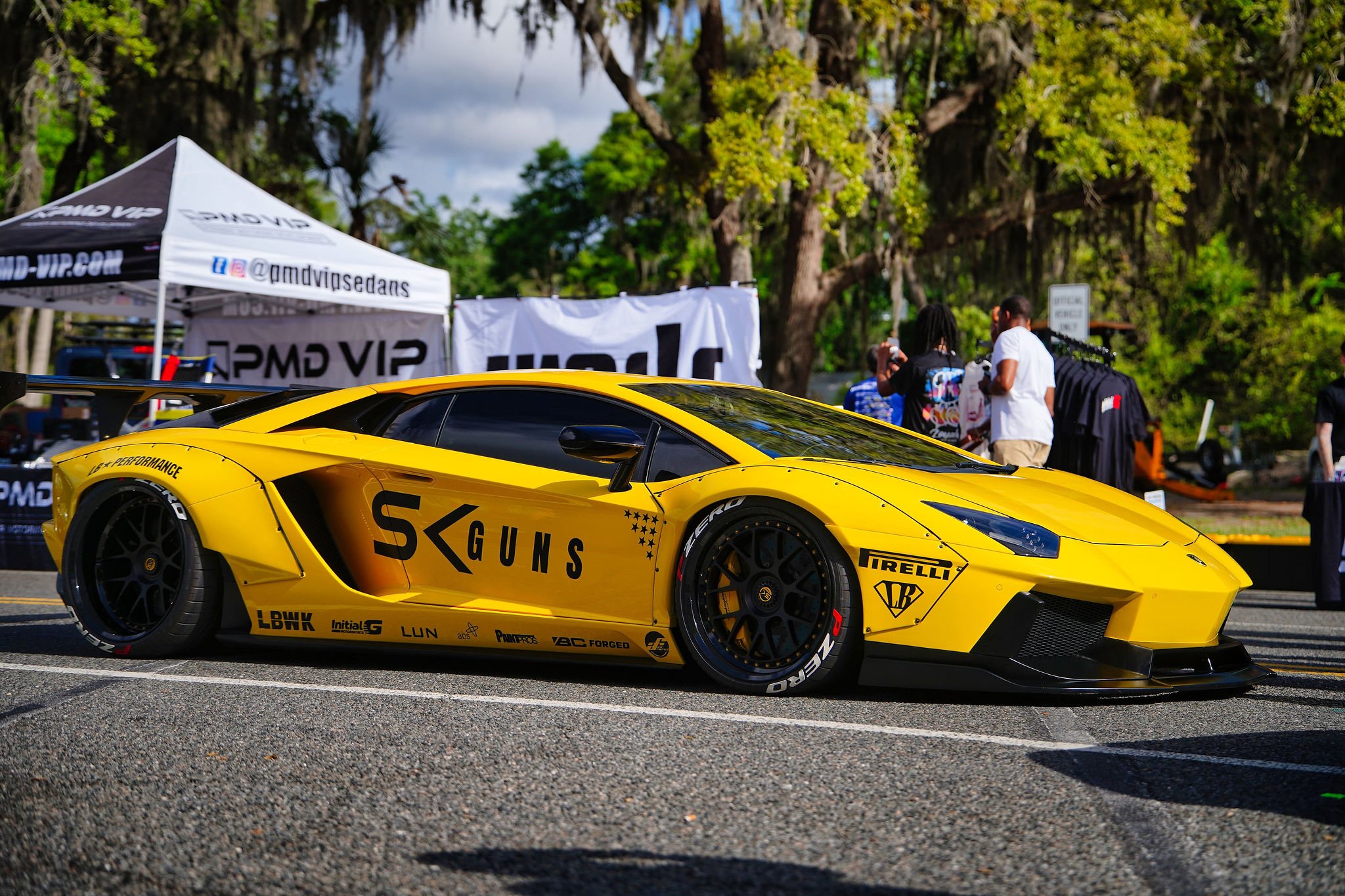 Yellow luxury sports car with black wheels parked at an outdoor event, surrounded by green trees and tents, with people taking photos in the background.