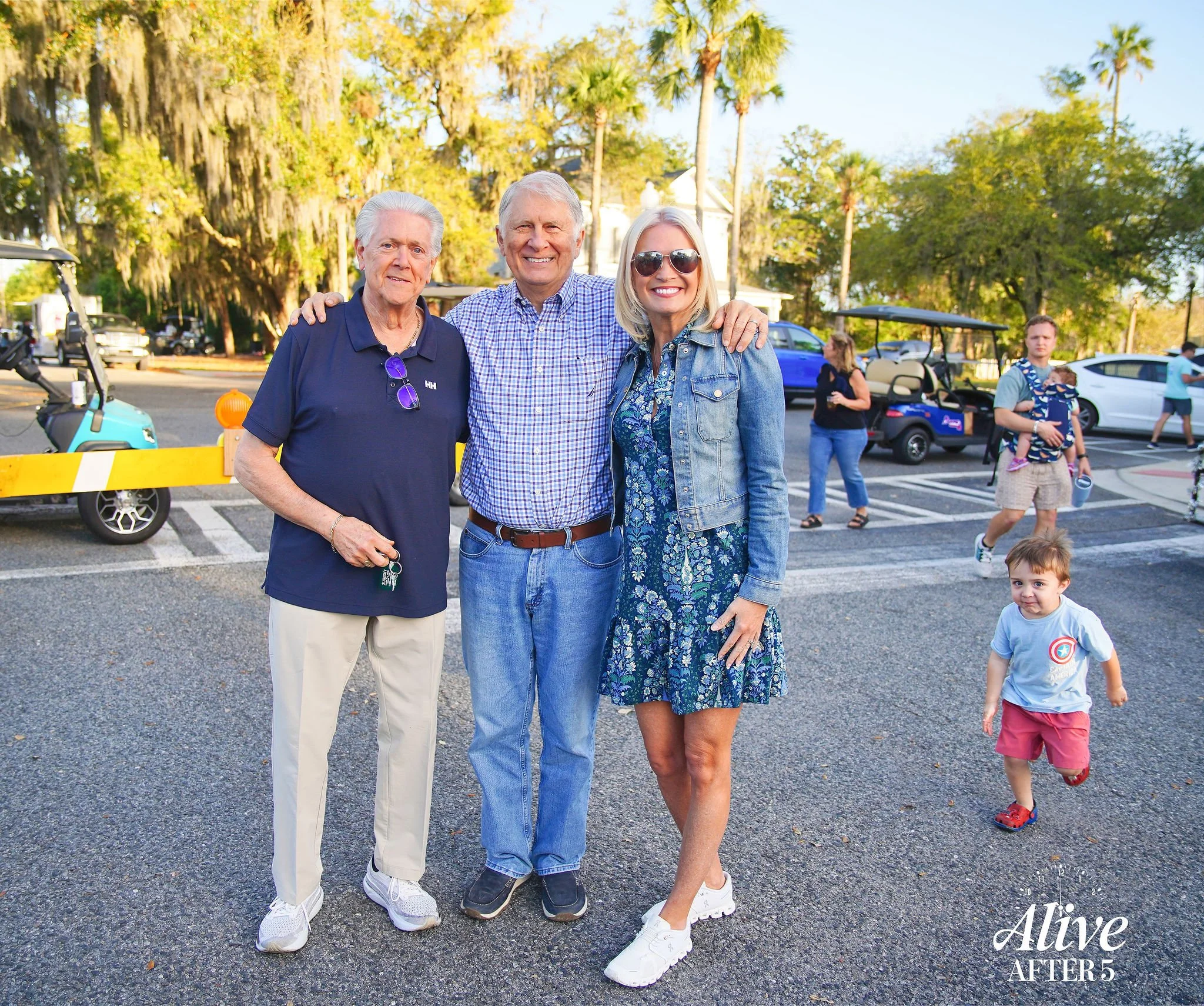 Three older adults and a young woman standing together in a parking lot with children and golf carts in the background. The group is smiling, and the scene appears to be outdoors on a sunny day.